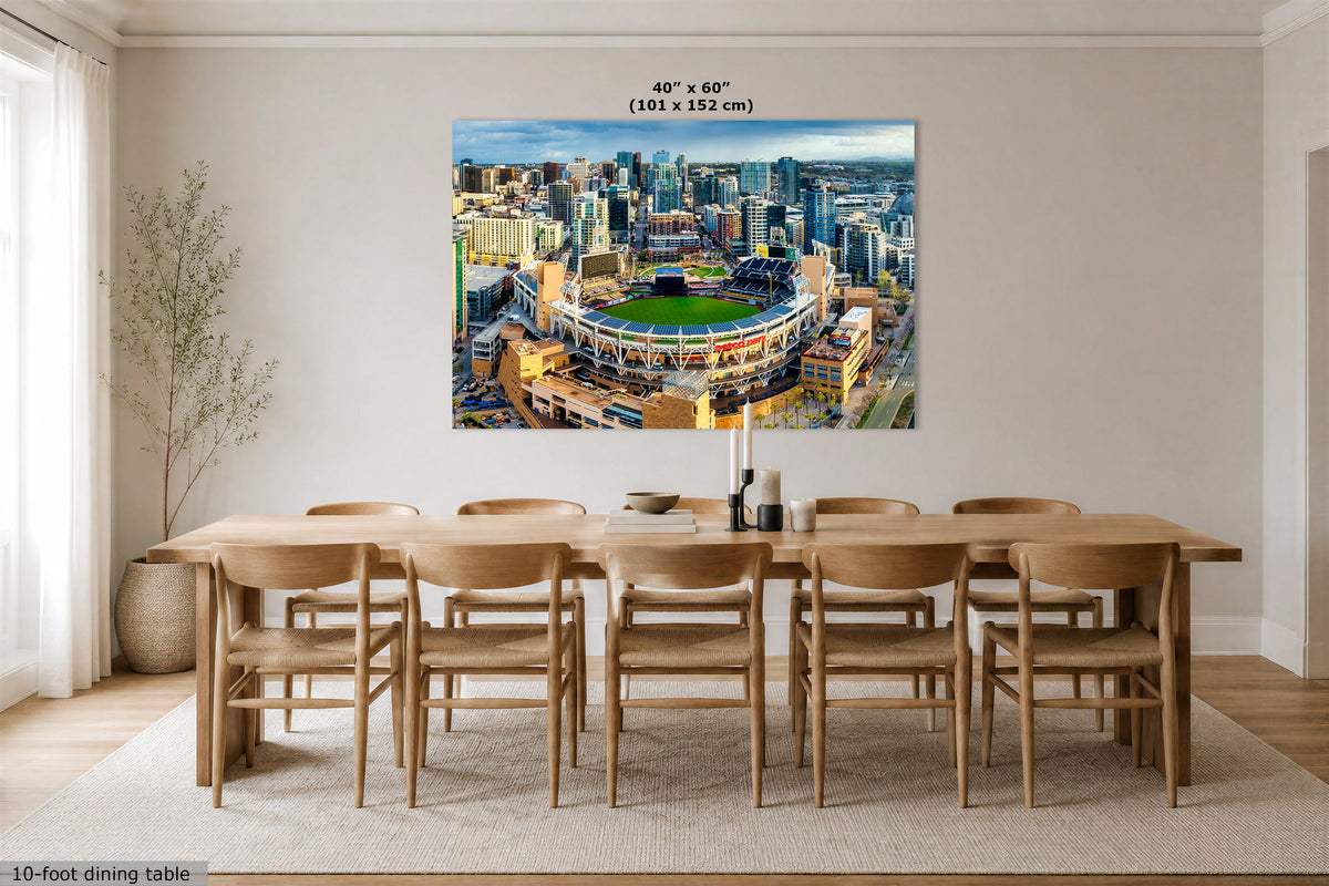 A large wall art print showing an aerial view of a stadium and city skyline, displayed above a wooden dining table in a bright modern dining room. Text at the top reads 40" x 60" (101 x 152 cm), and text at the bottom left says 10-foot dining table.
