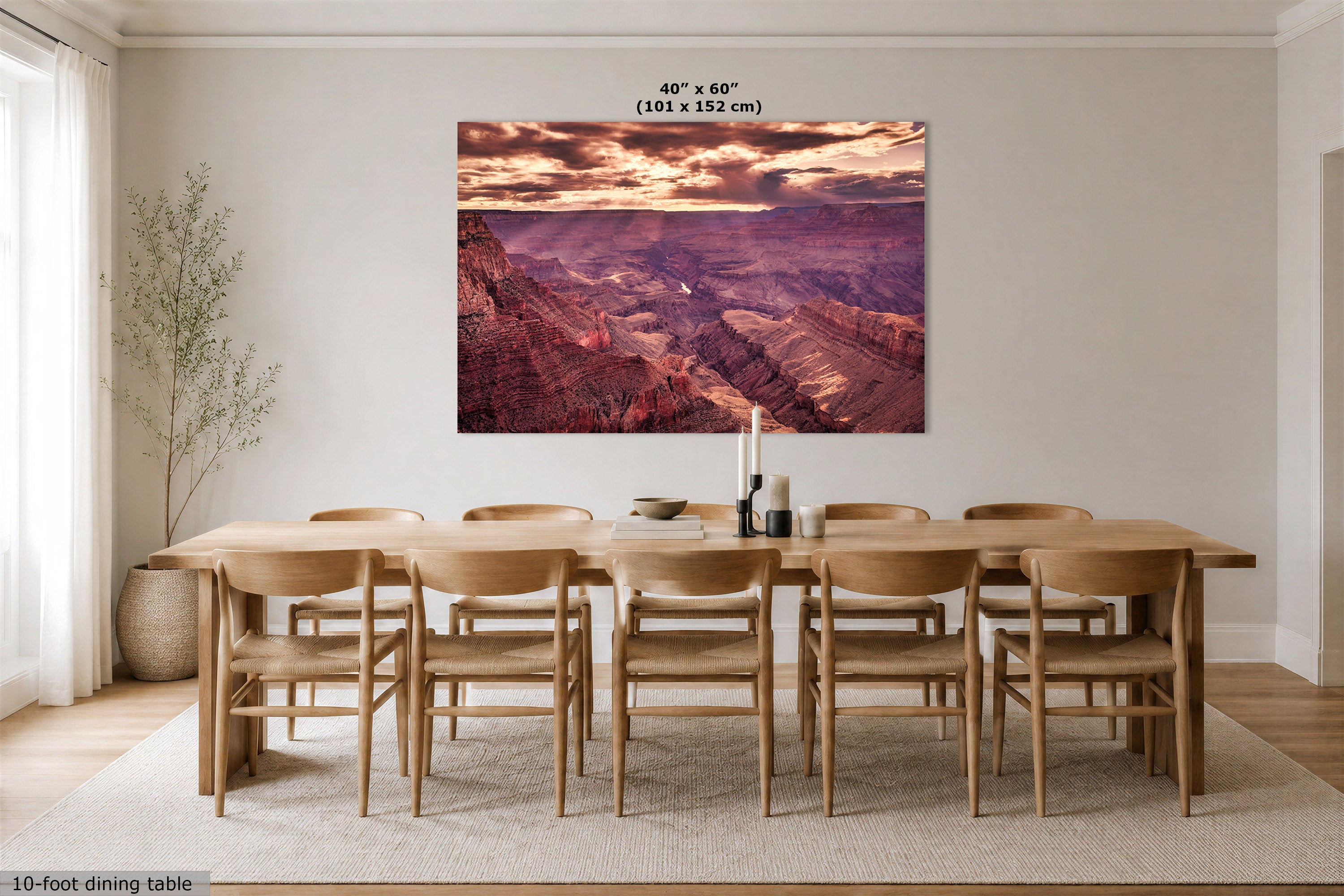A large rectangular wall art print showing a canyon landscape in warm sunset tones, displayed above a wooden dining table and chairs in a bright dining room. Text at the top reads 40 x 60" (101 x 152 cm), and a small note at the bottom left says 10-foot dining table.
