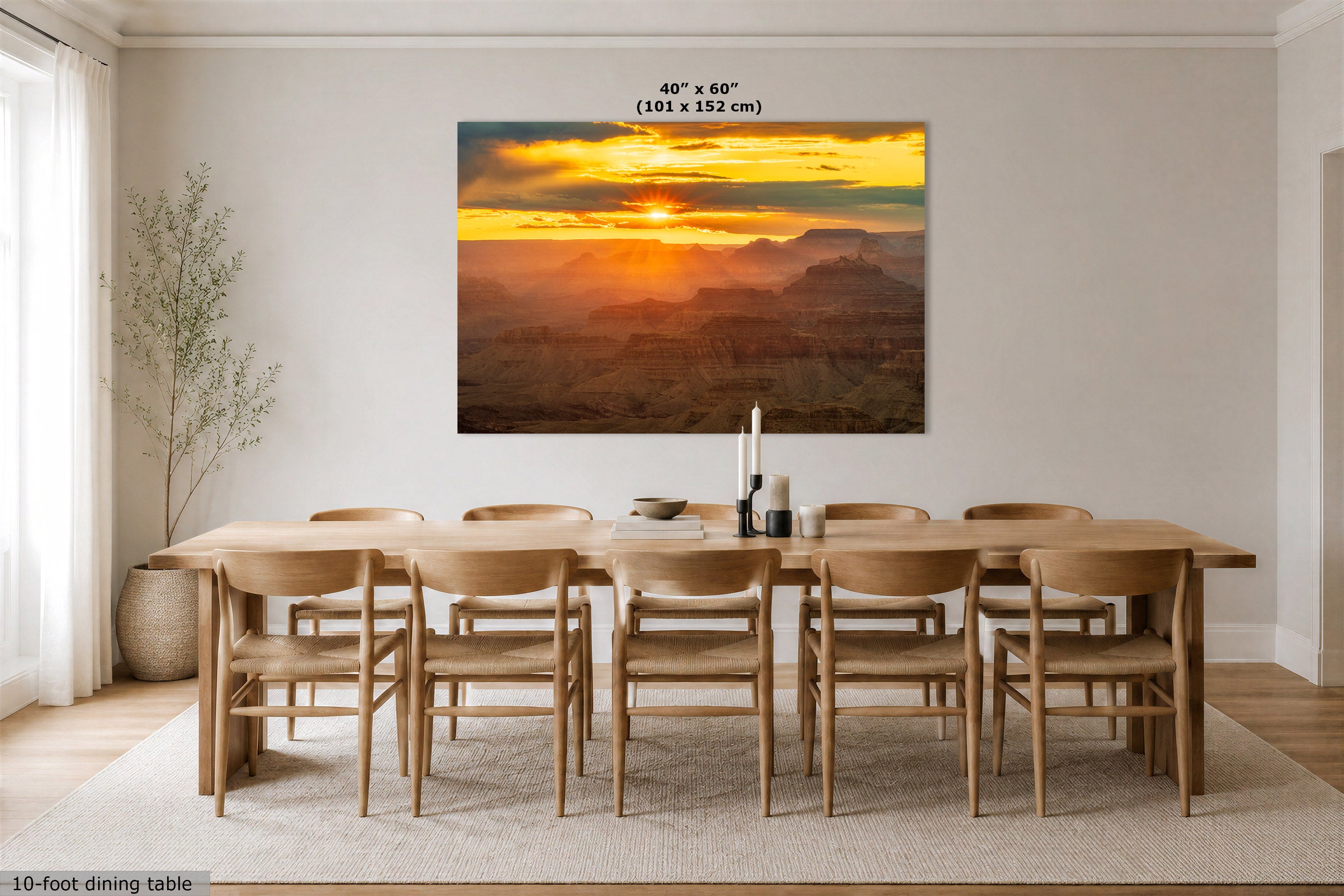 A large rectangular wall art print showing a dramatic sunset over layered canyon mountains, displayed above a wooden dining table and chairs in a bright neutral dining room. Text at the top of the print reads 40" x 60" (101 x 152 cm).