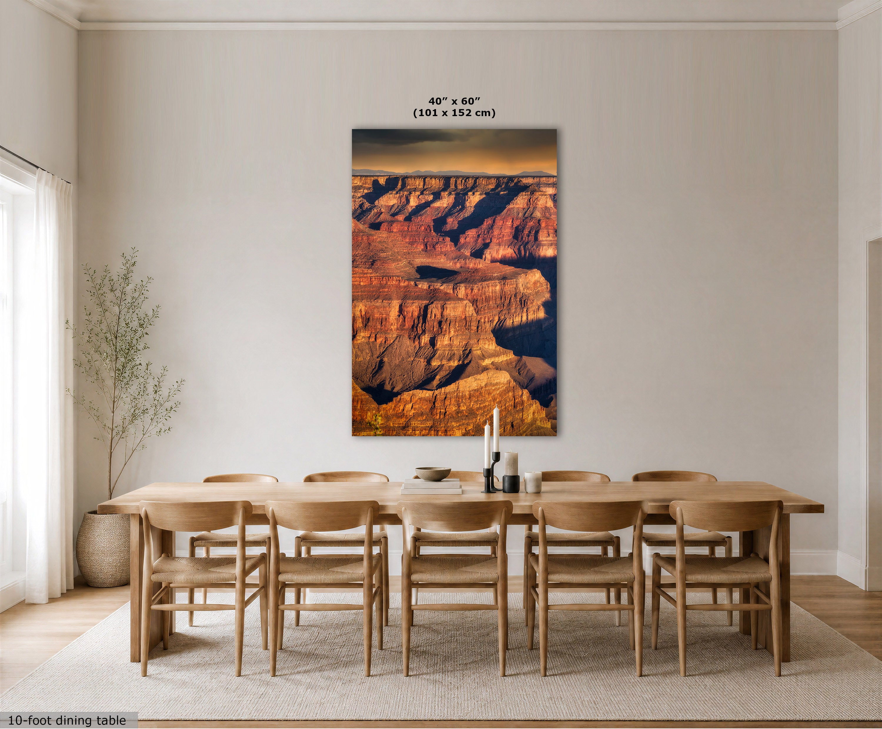 A large vertical wall art print showing a canyon landscape in warm orange, brown, and blue tones, displayed above a wooden dining table and chairs in a bright neutral dining room. Text at the top reads 40" x 60" (101 x 152 cm), and a small note at bottom left says 10-foot dining table.