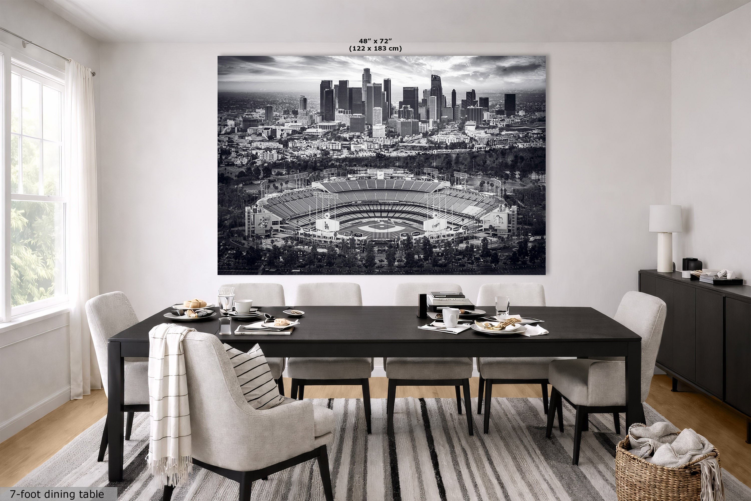 A large black and white wall art print of the Los Angeles skyline and Dodger Stadium, shown hanging in a modern dining room setting above a dark wood table and chairs.