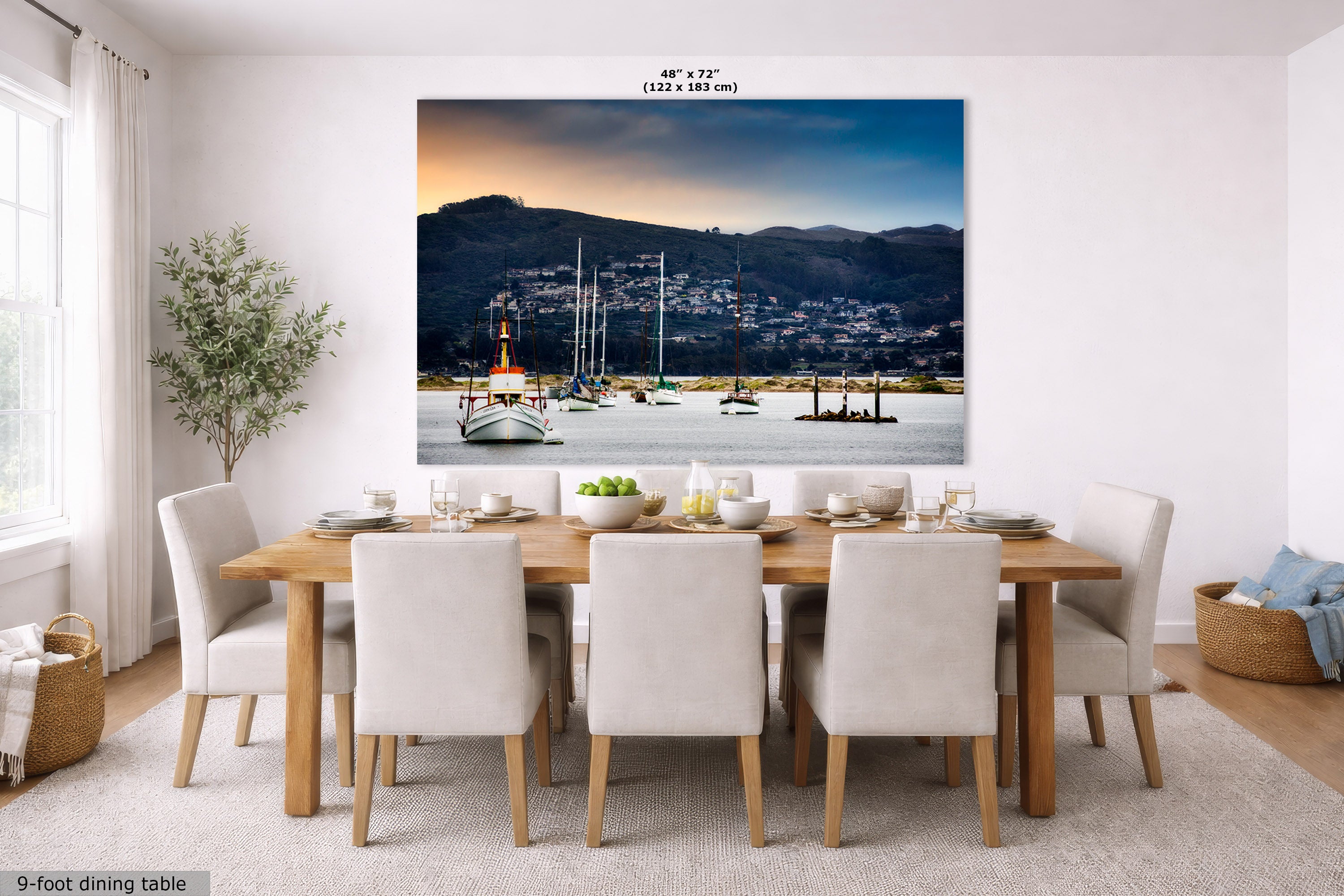 A large wall mural or canvas print depicting a coastal harbor scene with boats, mountains, and a sunset, displayed above a dining table in a bright modern room. The print has text at the top reading 48" x 72" and a small label at bottom left reading 9-foot dining table.