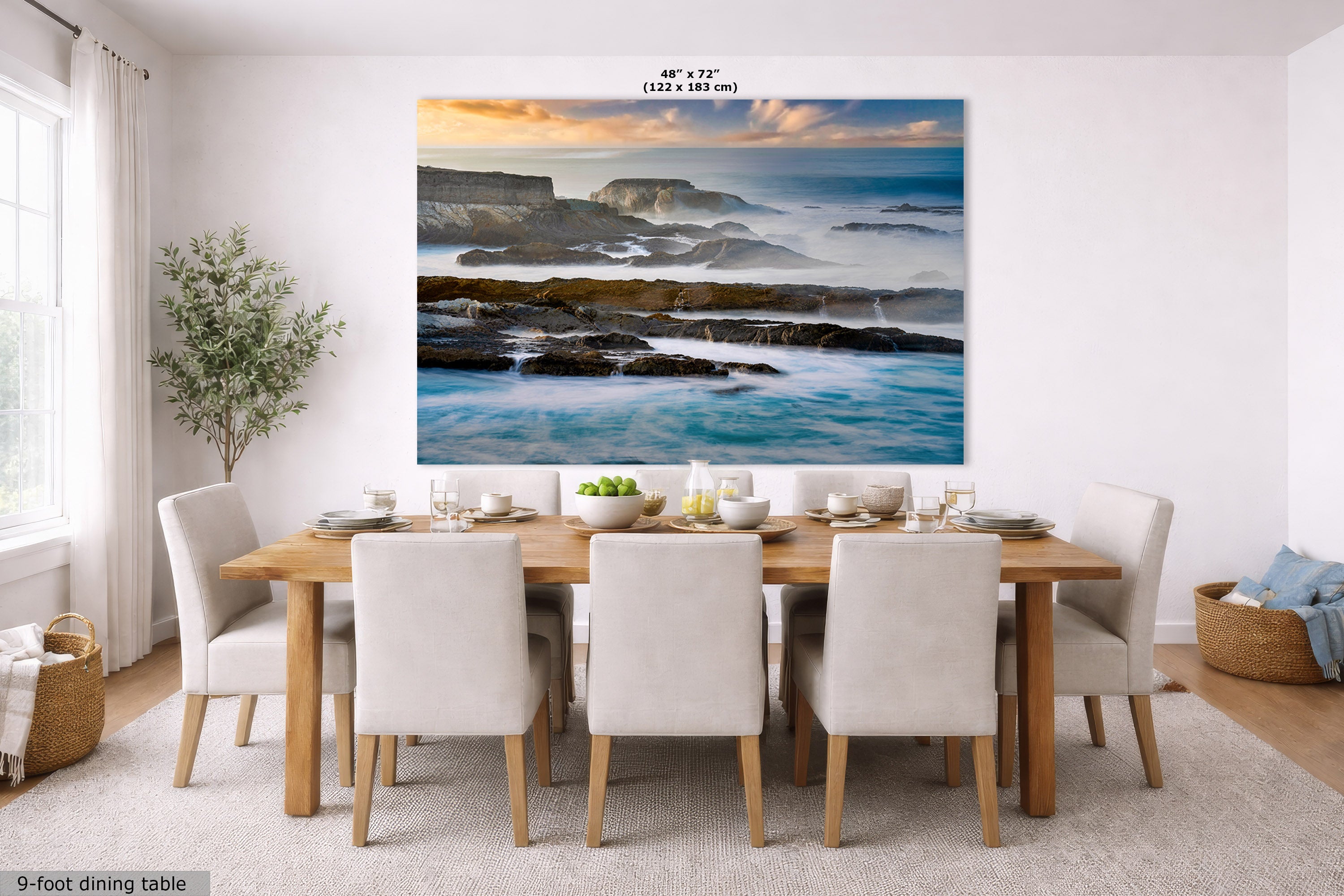 A large wall art print depicting a rocky coastal ocean scene with blue water, dark rocks, and a warm sunset sky, displayed above a dining table in a bright modern room. Text at the top reads 48" x 72" (122 x 183 cm), and a small label at bottom left says 9-foot dining table.