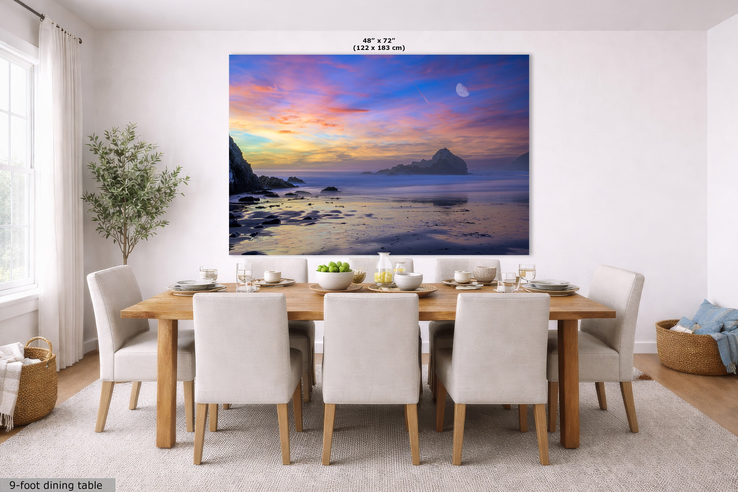 A large wall art print showing a colorful sunset over water with rocky cliffs and a crescent moon, displayed above a dining table in a bright modern room. The print includes size text at the top and a small label at bottom left reading 9-foot dining table.