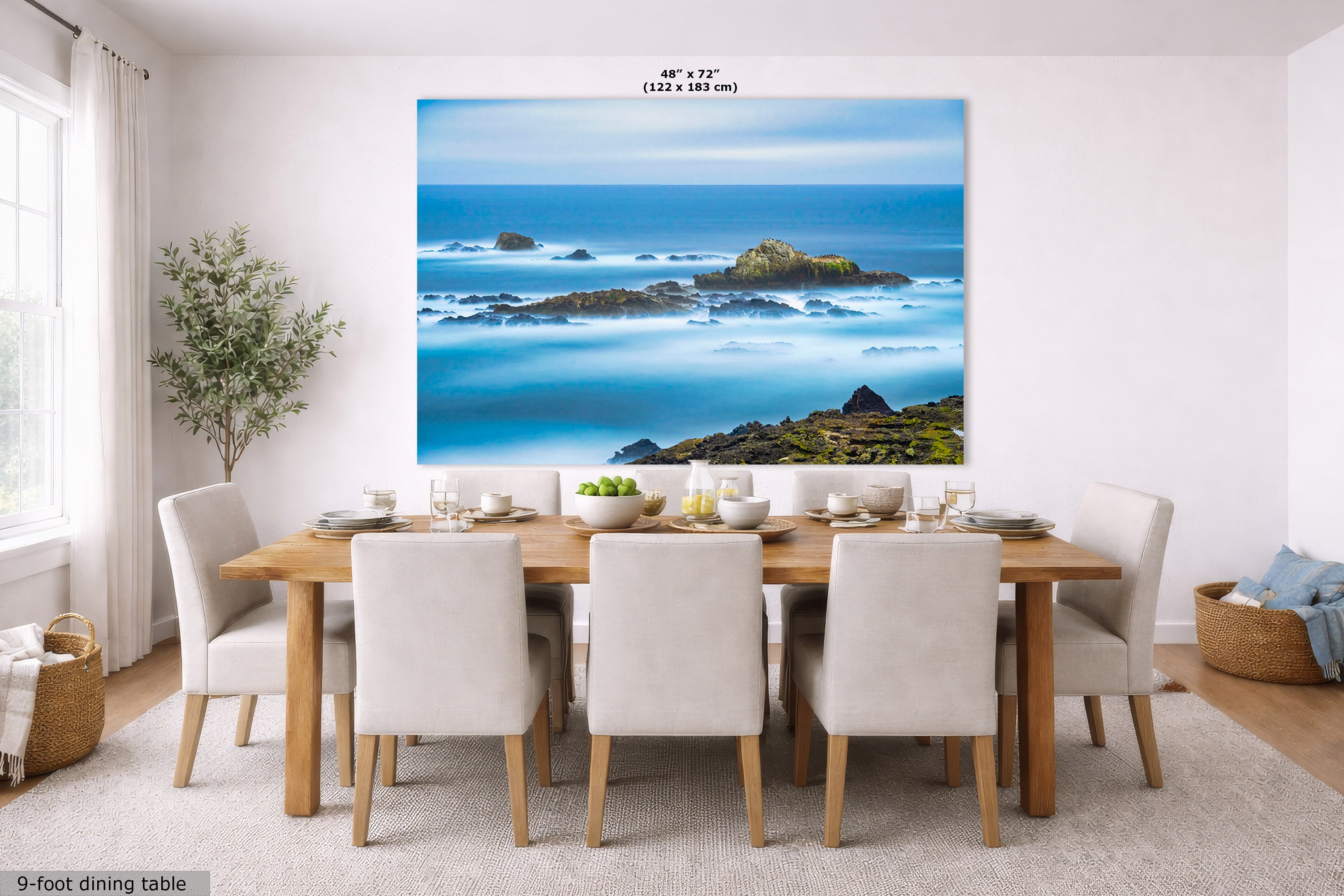 A large wall art print of a coastal rocky island landscape in blue and turquoise tones, displayed above a dining table in a bright modern room. The print includes size text at the top and a small label at bottom left reading 9-foot dining table.