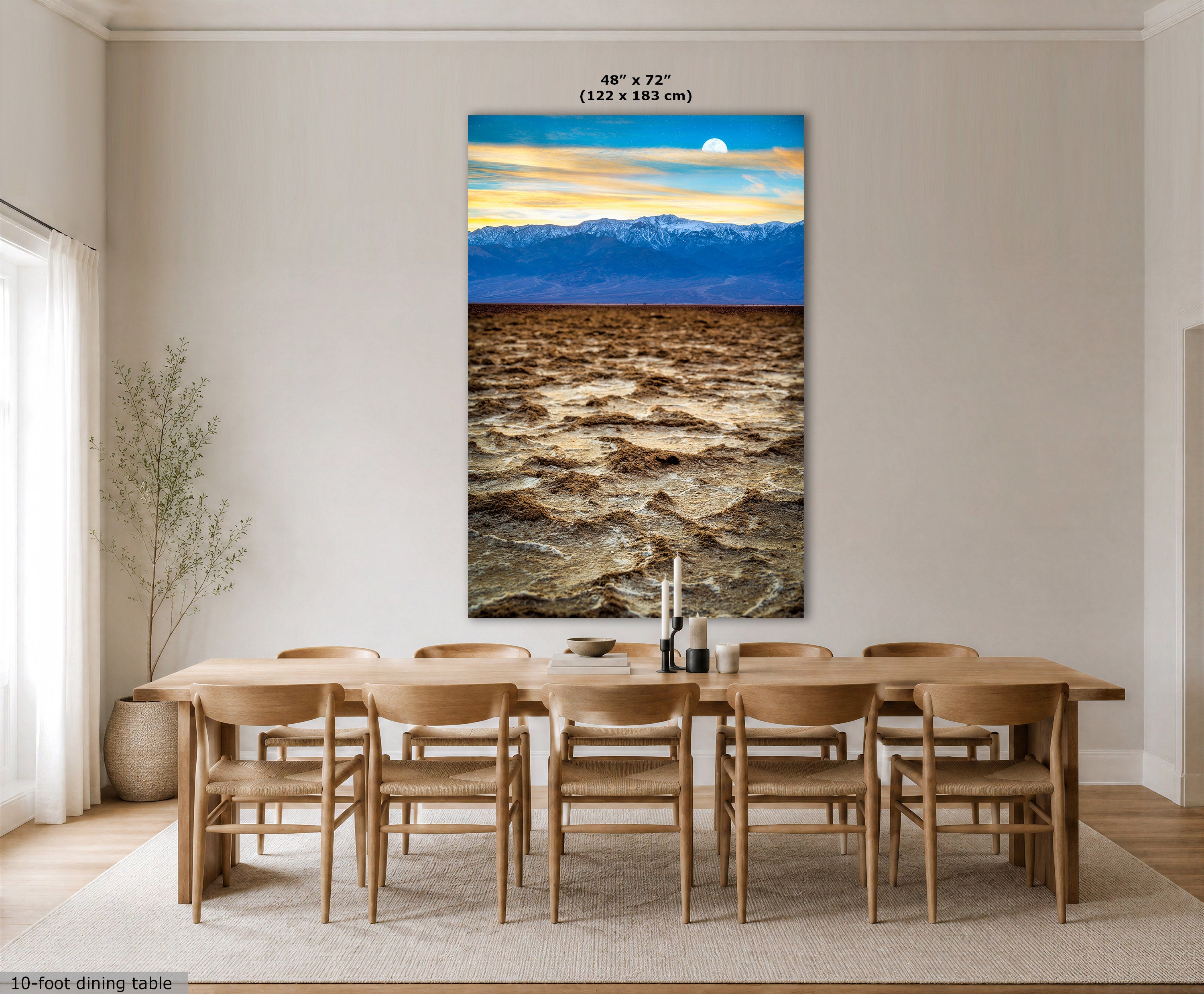 A large vertical wall art print showing a dramatic mountain landscape with blue peaks and a golden sky, displayed above a wooden dining table and chairs in a bright modern dining room. The print includes size text at the top and a small label at the bottom left.