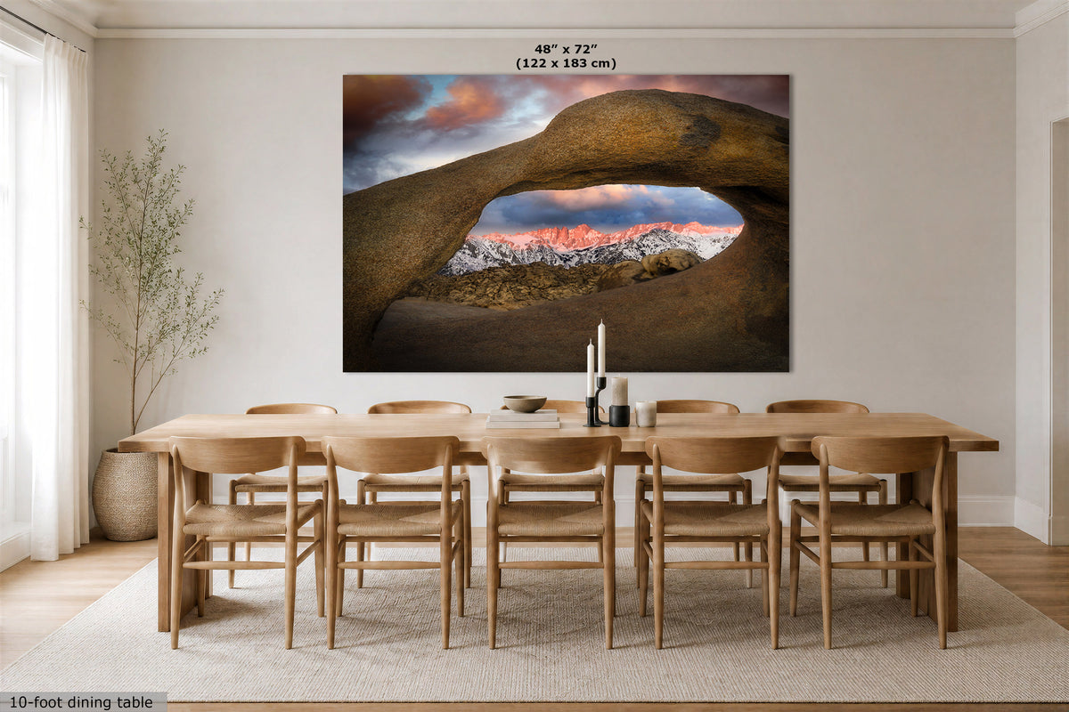 A large wall art print viewing through the Mobius Arch at the Alabama Hills, looking at the Sierra Nevada mountains at sunrise, displayed above a wooden dining table and chairs in a bright dining room. The print includes size text at the top and a small caption at the bottom left.