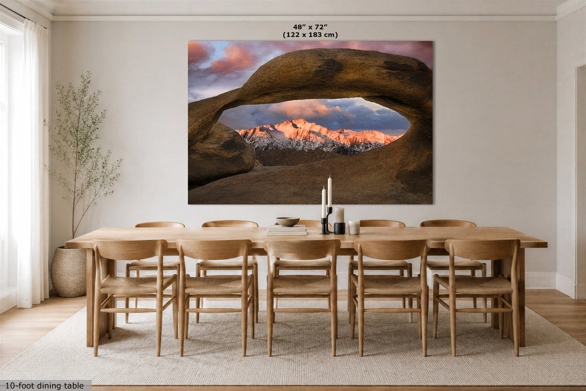 A large wall art print showing a desert rock arch framing snow-capped mountains at sunset, displayed above a wooden dining table and chairs in a bright neutral dining room. Text at the top reads 48" x 72" (122 x 183 cm), and a small note at bottom left says 10-foot dining table.