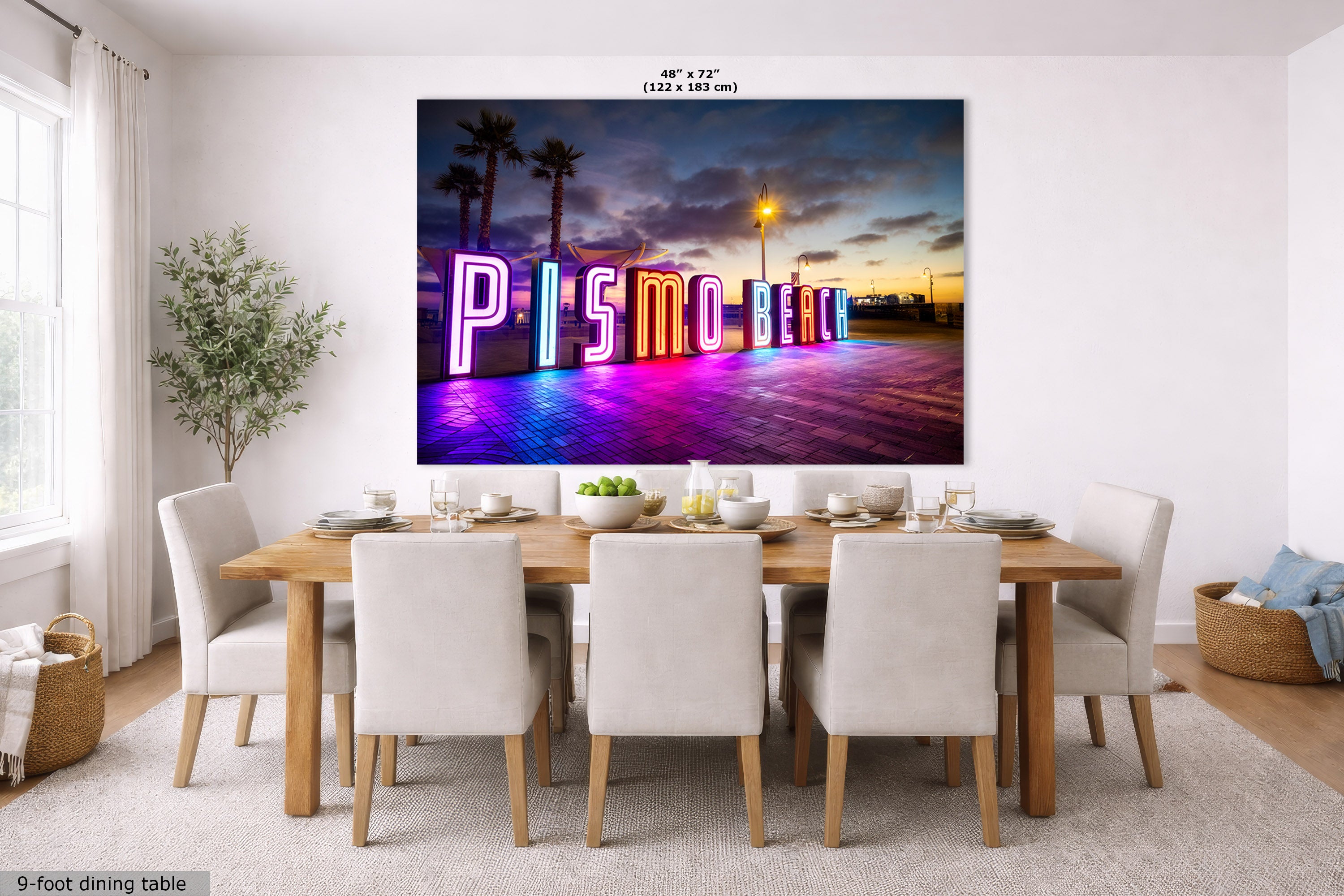 A large wall art print showing the neon 'Pismo Beach' sign at sunset, displayed above a dining table in a bright modern room. The print includes size text at the top and a small label at bottom left reading '9-foot dining table'.