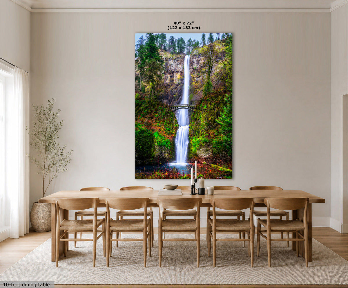 A large vertical wall art print showing a waterfall in a forest, displayed above a wooden dining table and chairs in a bright, modern dining room. The print includes size text at the top and a small label at the bottom left.