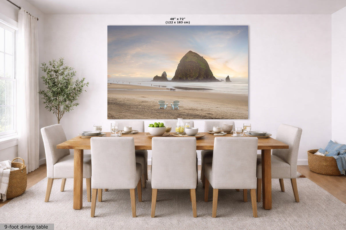 A large wall art print depicting Haystack Rock on a beach at sunset, displayed in a dining room above a wooden dining table and six upholstered chairs. The print has a small size label at the top reading 48" x 72" (122 x 183 cm).