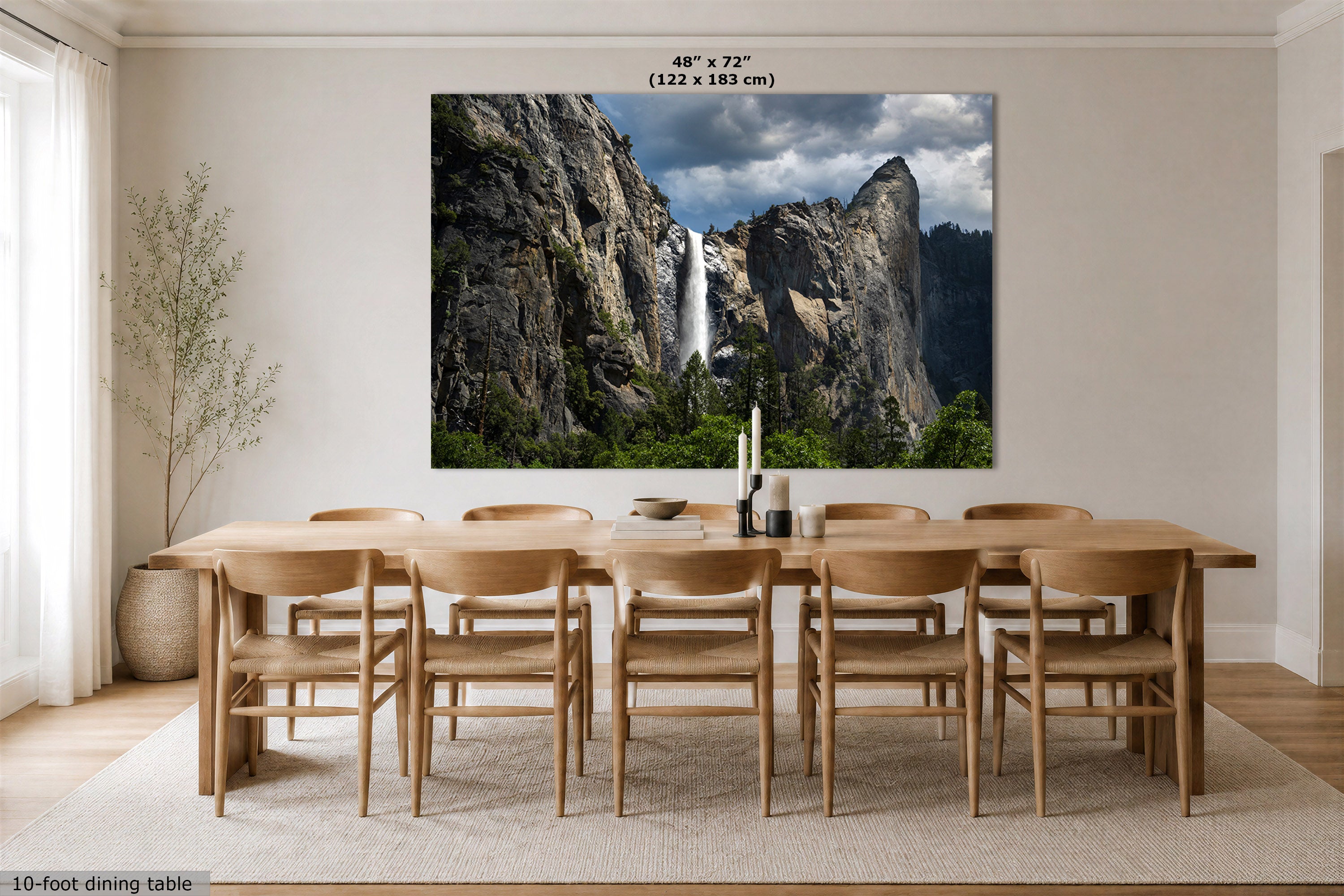 A large wall art print depicting a dramatic waterfall between rocky cliffs and green trees, displayed above a wooden dining table and chairs in a bright, modern dining room. The print includes size text at the top reading 48" x 72" (122 x 183 cm).