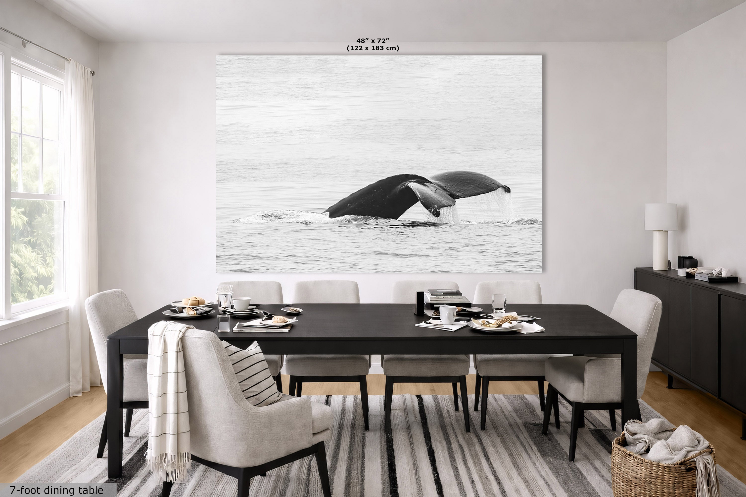 A large black and white whale wall art print hangs above a dark dining table and light upholstered chairs in a bright modern dining room. The print depicts a whale tail breaking the ocean surface.