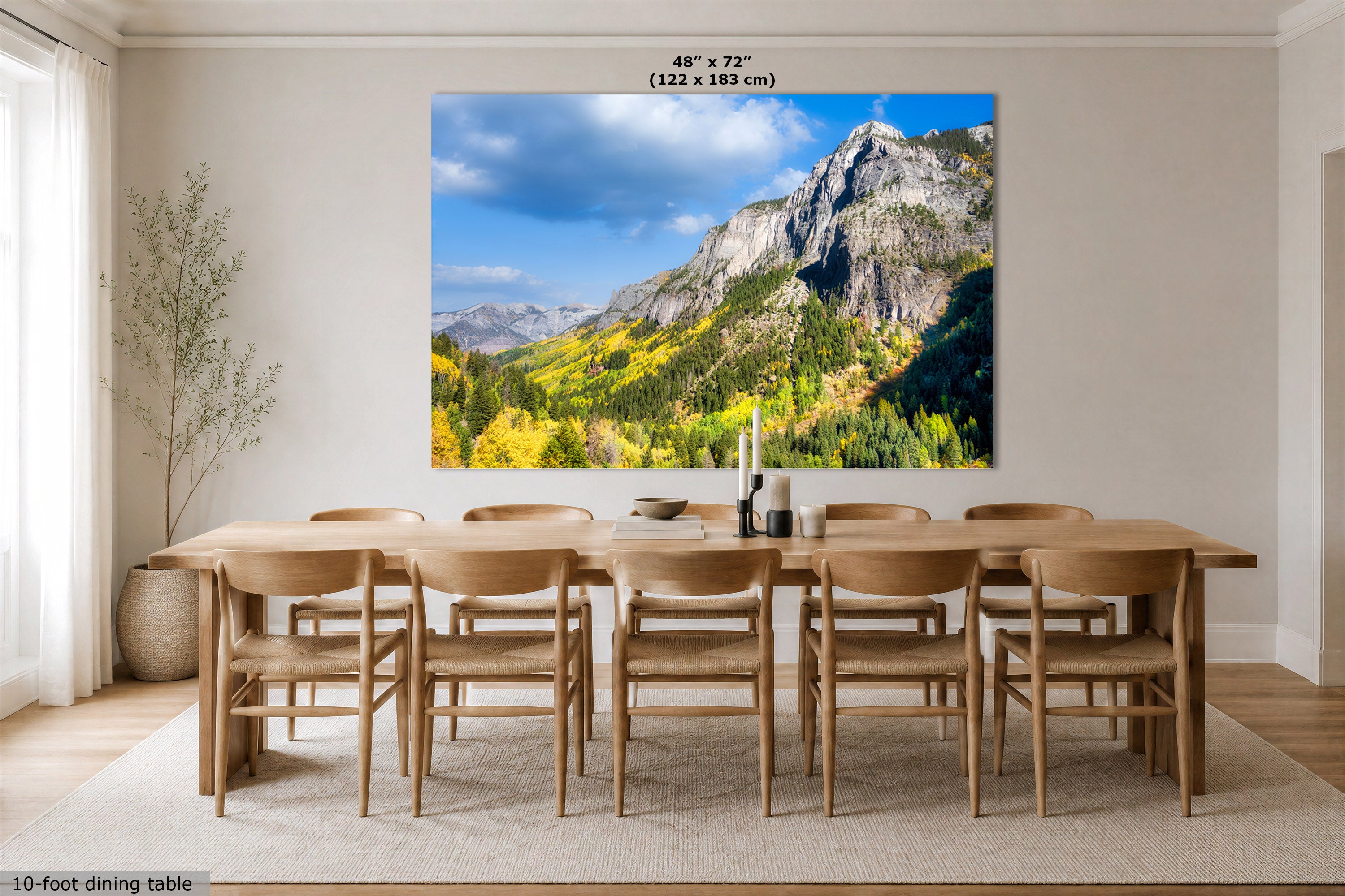 A large wall art print showing a mountain landscape with blue sky and autumn foliage, displayed above a wooden dining table and chairs in a bright dining room. Text at the top reads 48" x 72" (122 x 183 cm).