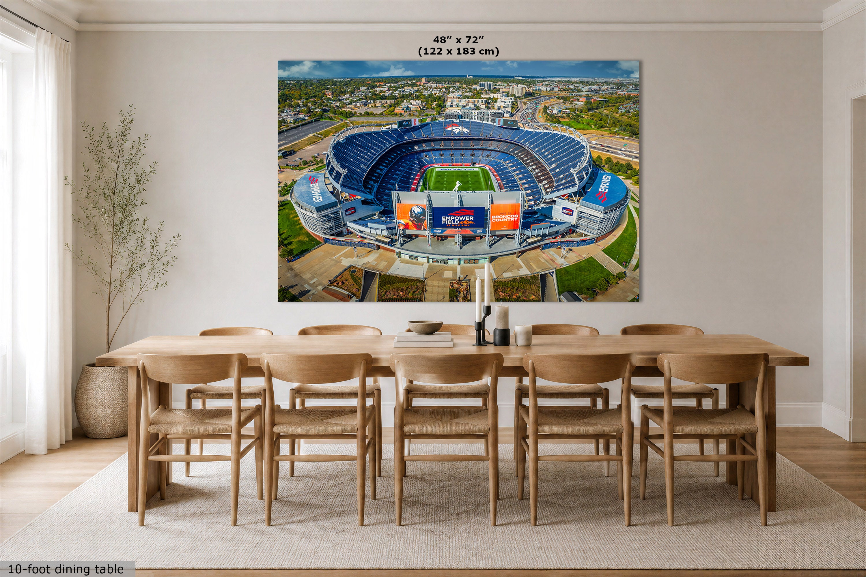 A large rectangular wall art print showing an aerial view of a blue stadium, displayed above a wooden dining table in a bright modern room. The print includes size text at the top reading 48" x 72" (122 x 183 cm).