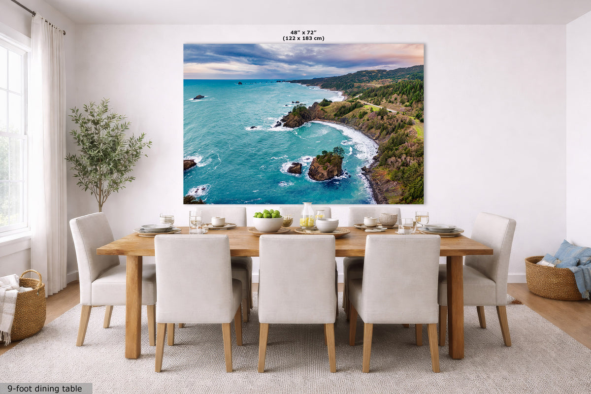 A large wall art print showing a coastal landscape with turquoise ocean, rocky shoreline, and green hills, displayed above a dining table in a bright modern room. Small text at the top reads 48" x 72" (122 x 183 in), and a label at bottom left says 9-foot dining table.