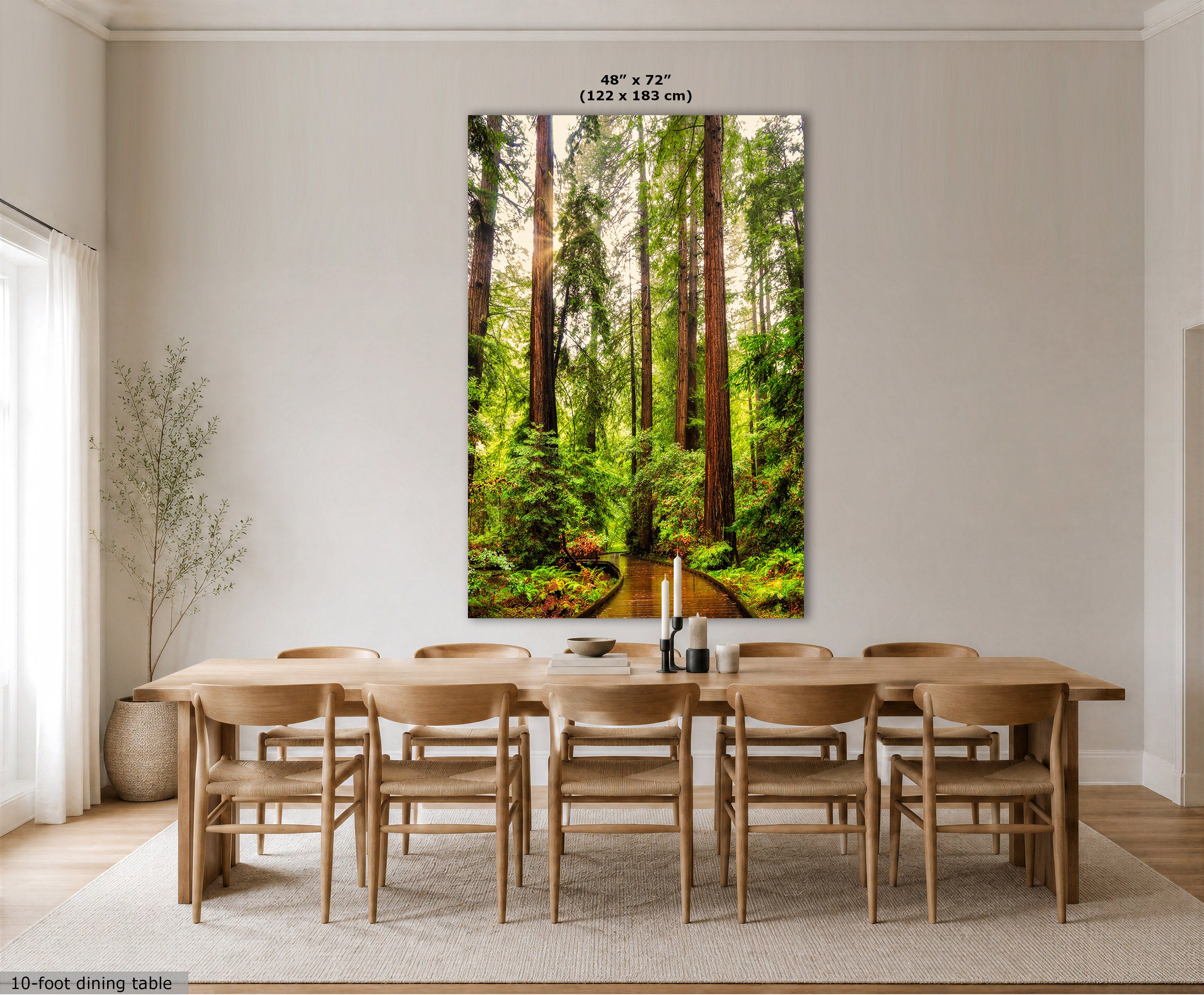 A large vertical wall art print showing a sunlit redwood forest path, displayed above a light wood dining table and chairs in a bright neutral dining room. The print includes size text at the top and a small caption at the bottom left.