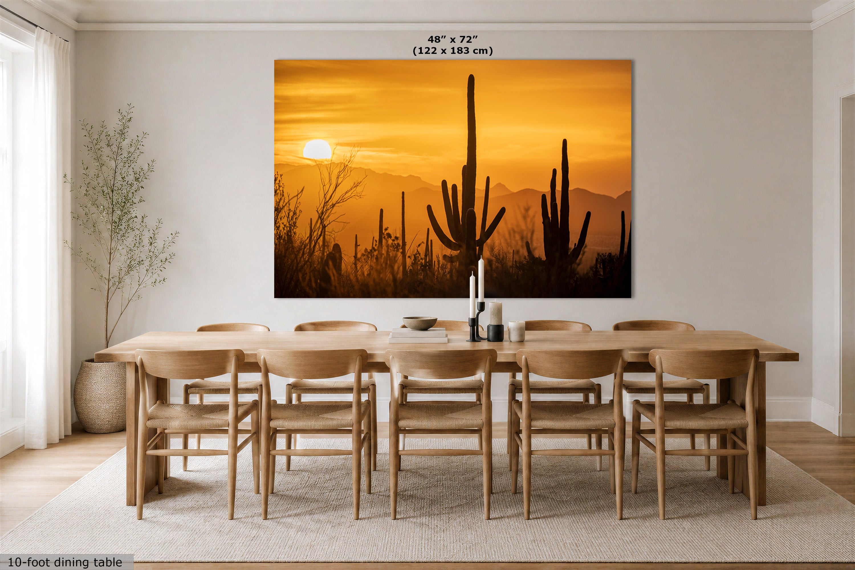 A large rectangular wall art print showing a desert sunset with cacti and mountains, displayed above a wooden dining table and chairs in a bright dining room. The print includes size text at the top reading 48" x 72" (122 x 183 cm).