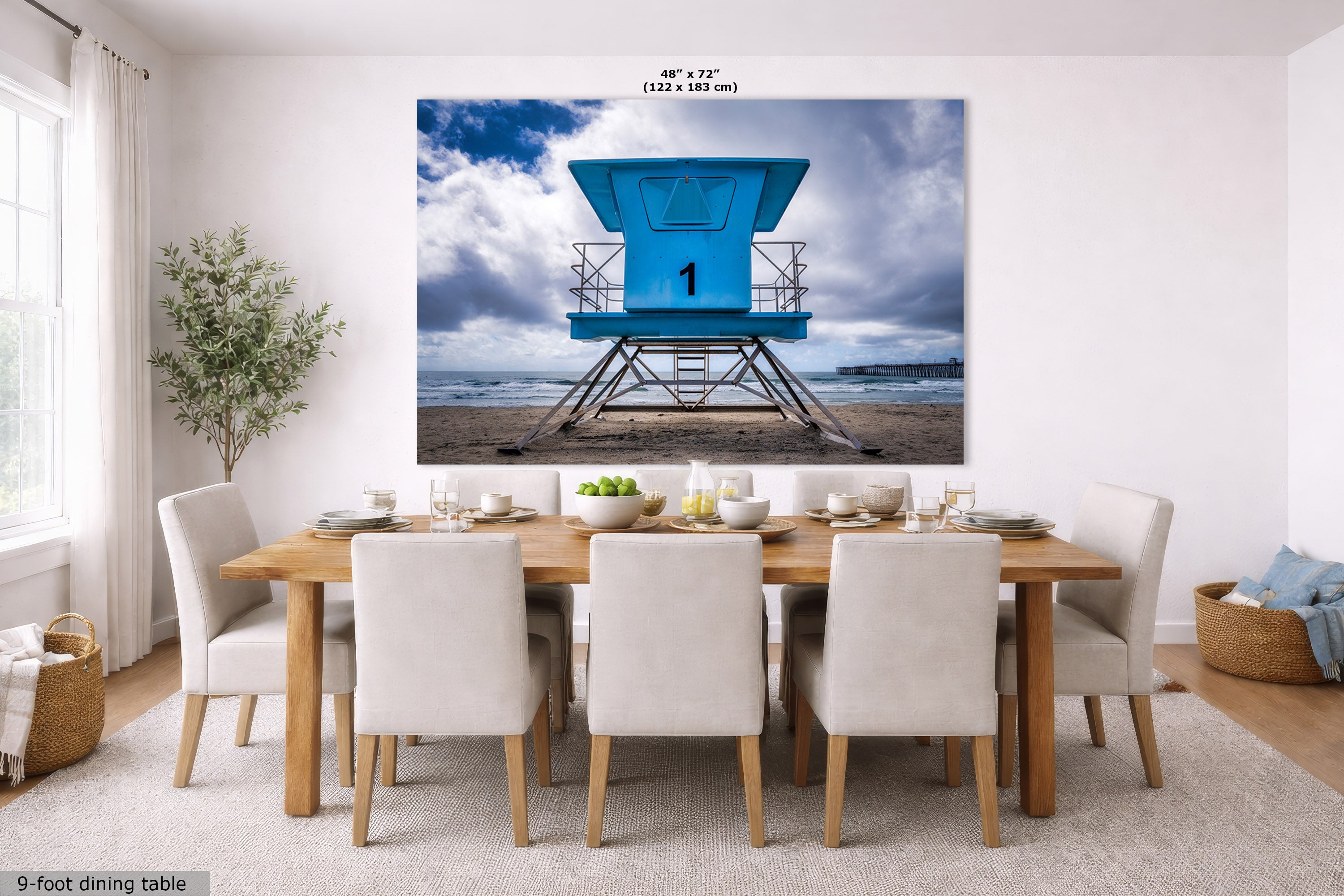 A large wall art print showing a bright blue lifeguard tower on a beach under a cloudy sky, displayed above a dining table in a modern white dining room. The print includes size text at the top and a small label at the bottom left.