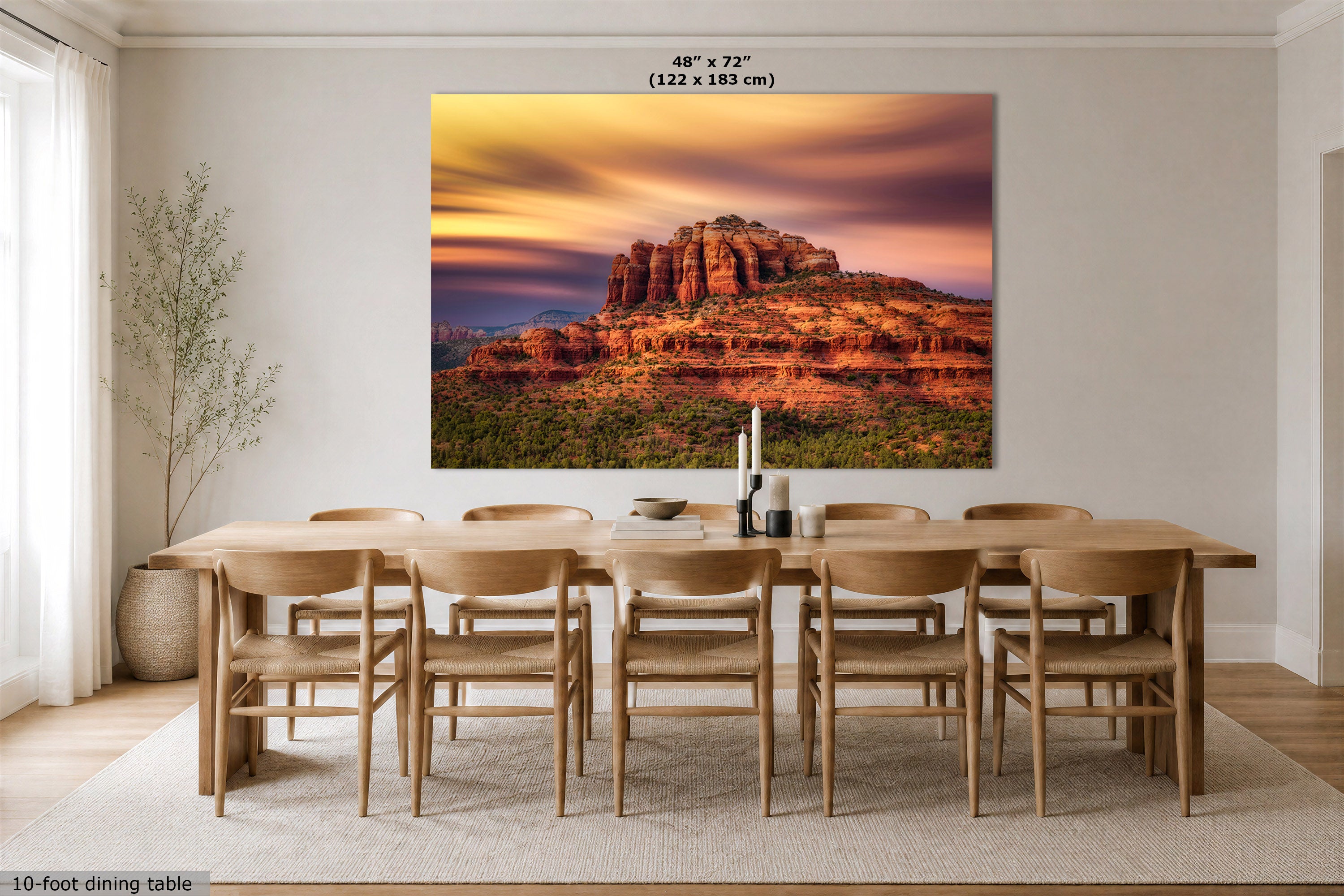 A large wall art print showing a desert rock formation at sunset, displayed above a wooden dining table and chairs in a bright dining room. The print includes size text at the top and a small note at bottom left.