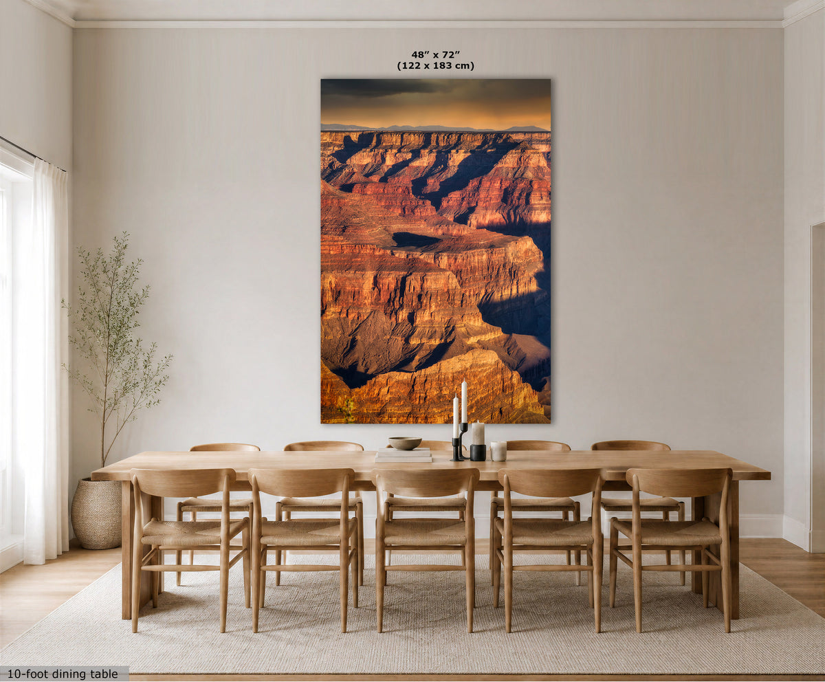 A large vertical wall art print depicting the Grand Canyon in warm sunset tones, displayed above a wooden dining table and chairs in a bright, modern dining room. The print includes size text at the top and a small label at the bottom left.