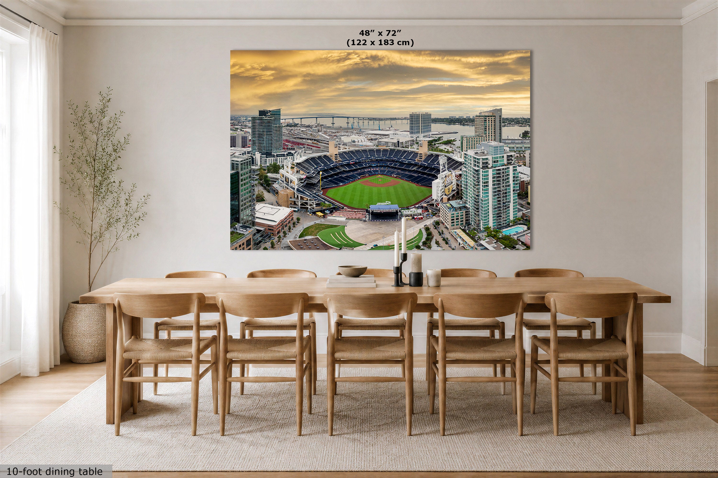 A large rectangular wall art print showing an aerial view of a baseball stadium at sunset, displayed above a wooden dining table and chairs in a bright modern dining room. The print includes size text at the top reading 48" x 72" (122 x 183 cm).