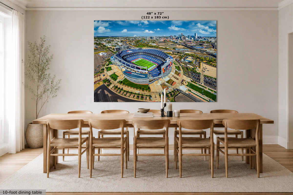 An Empower Field Denver skyline panorama wall art print showing an aerial view of the Broncos' Mile HIgh Stadium under a cloudy sky, displayed above a wooden dining table and chairs in a bright modern room. Text at the top of the print reads 48" x 72" (122 x 183 cm).