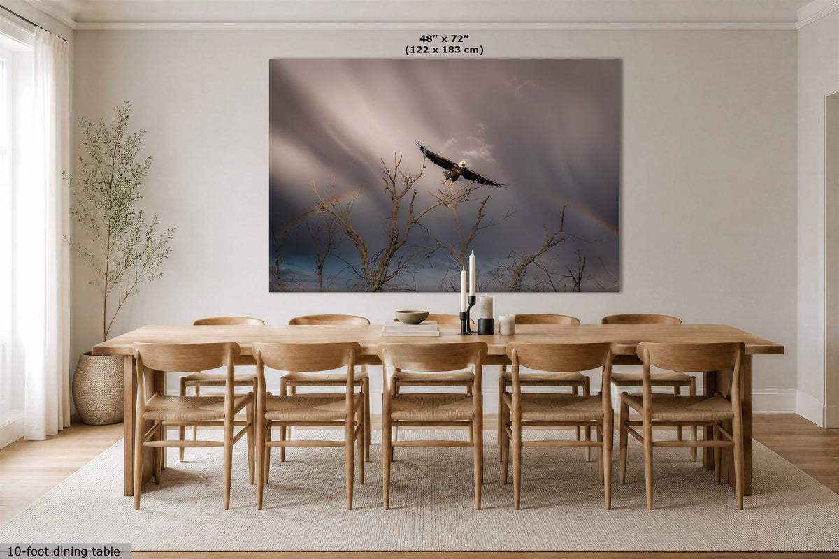 A large wall art print showing a bald eagle flying through dramatic storm clouds with a faint rainbow, displayed above a wooden dining table and chairs in a bright, modern dining room. The print includes size text at the top reading 48" x 72" (122 x 183 cm).