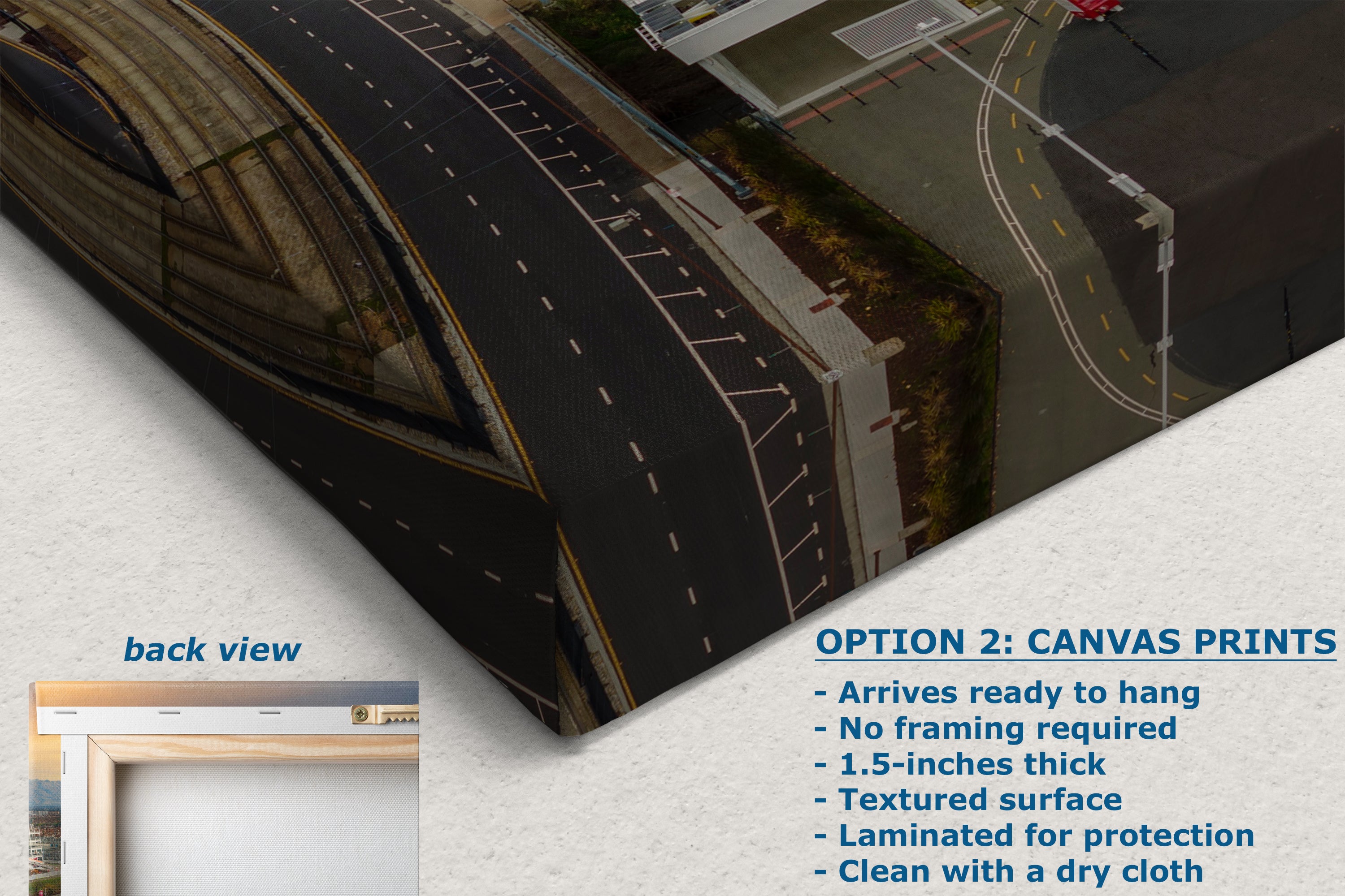 The image is a canvas print featuring an aerial view of a road with buildings and a bridge in the background. The text below the image provides information about the print, including that it is a back view of the print, arrives ready to hang, does not require framing, is 1.5-inches thick, has a textured surface, is laminated for protection, and can be cleaned with a dry cloth.