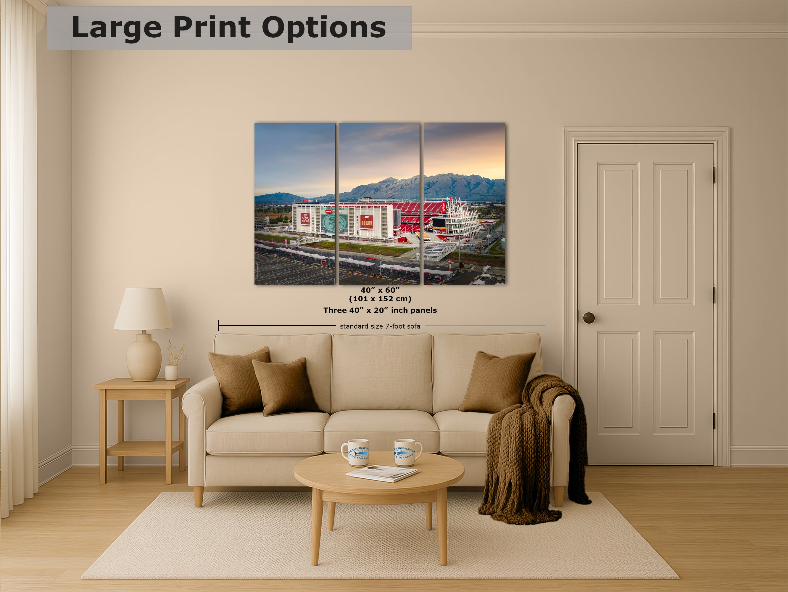 The image is a living room with a large, colorful painting of a stadium or sports arena hanging on the wall. The painting is divided into three panels, depicting the stadium from different angles. The room has a beige sofa, a coffee table, and a lamp, creating a cozy and inviting atmosphere.
