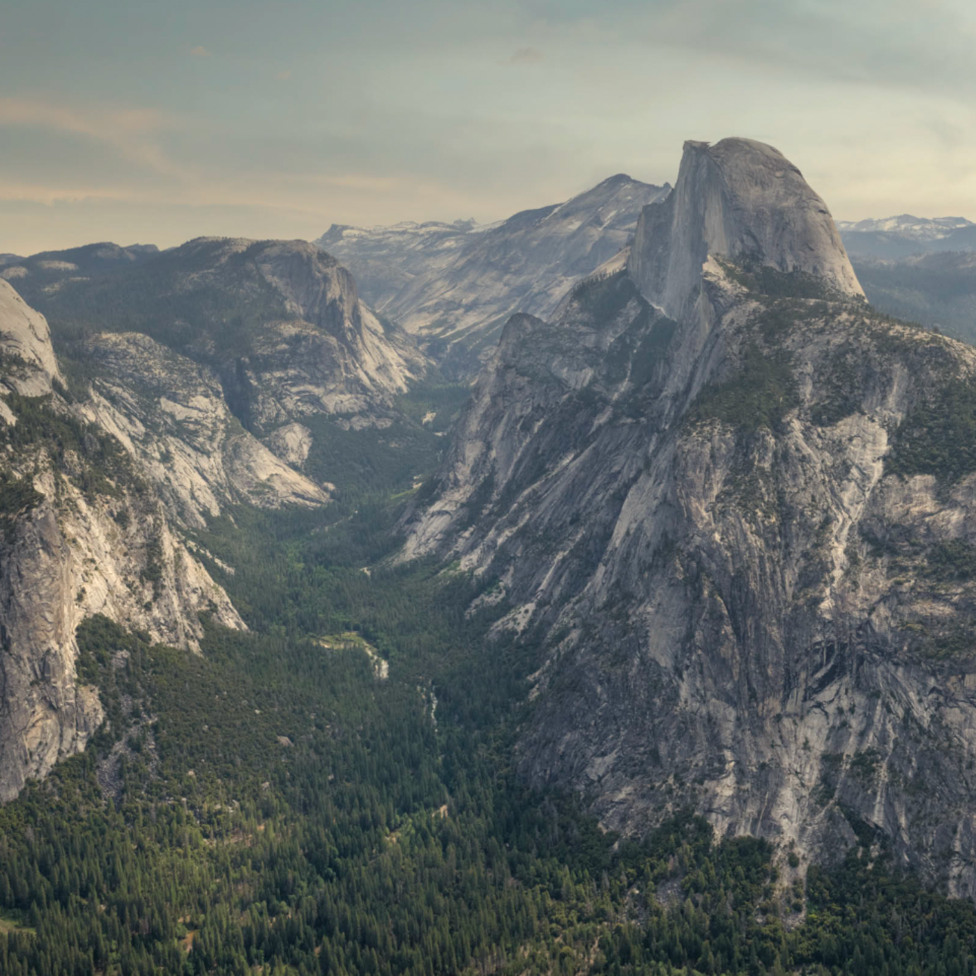 A scenic landscape photograph of a dramatic mountain range with large granite cliffs, a deep forested valley, and a hazy sky.