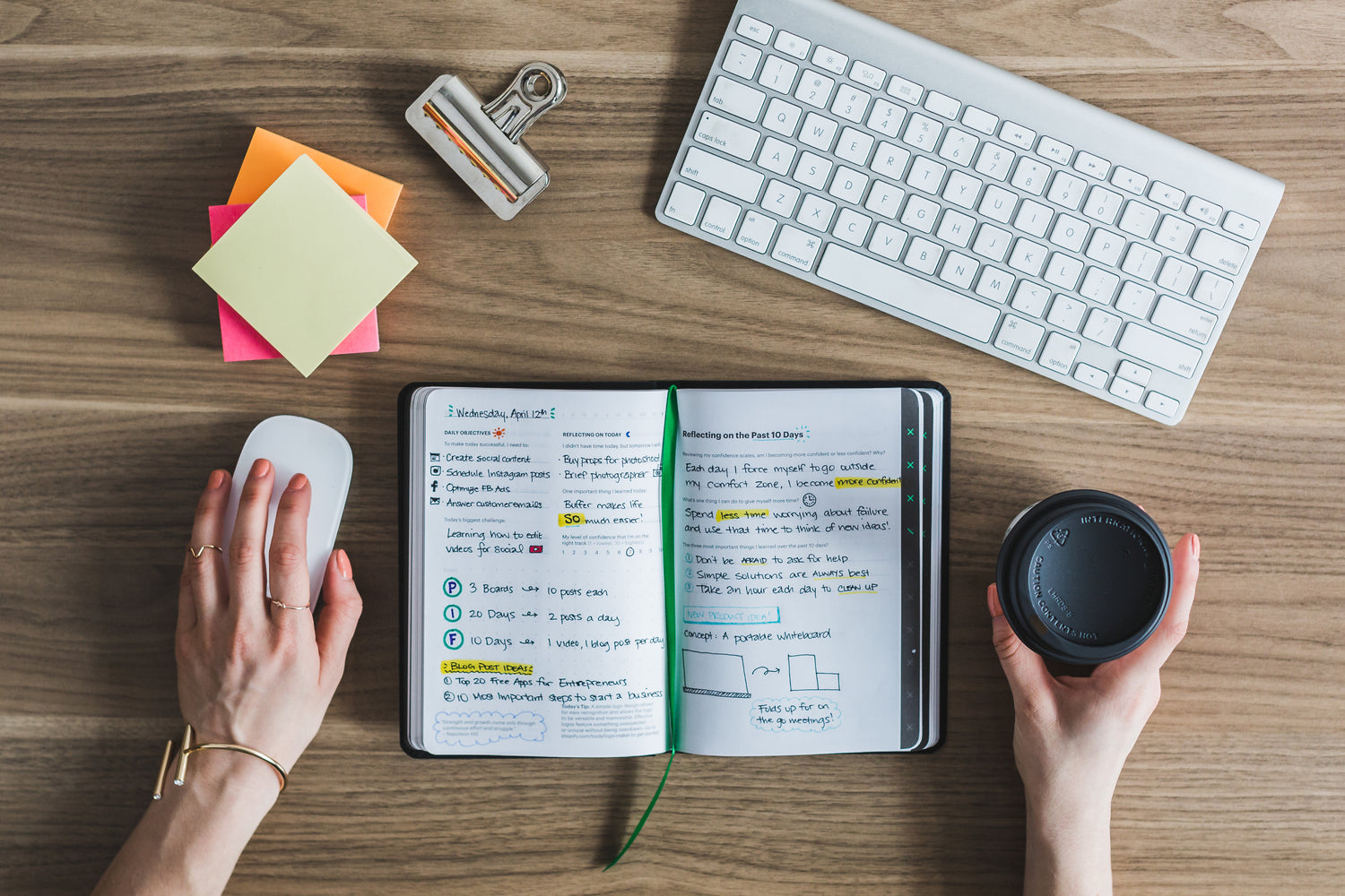 Overhead view of an over work-ed person's workspace featuring a person's hands using a mouse and holding a coffee cup, with a digital planner, keyboard, and sticky notes on a wooden desk