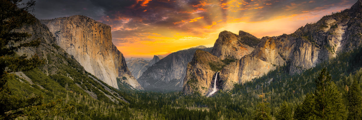 A dramatic landscape photograph of a mountain valley of Yosemite Valley from Tunnel View at sunrise, with steep rocky cliffs, evergreen forests, a waterfall, and a glowing orange and yellow sky.