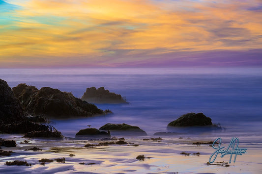 Serene sunset over Big Sur with smooth purple sand leading to calm waters, reflecting a tapestry of vibrant skies.