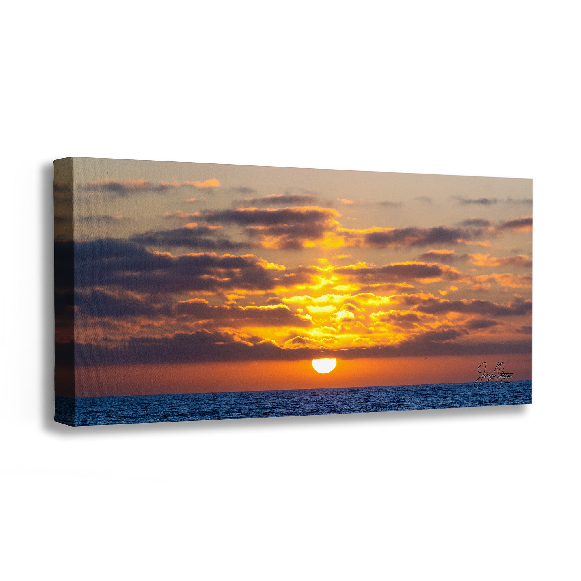 A rectangular canvas wall art print showing a dramatic sunset over the ocean, with glowing orange and yellow clouds, a bright sun near the horizon, and deep blue water. The canvas is photographed at an angle against a clean white background.
