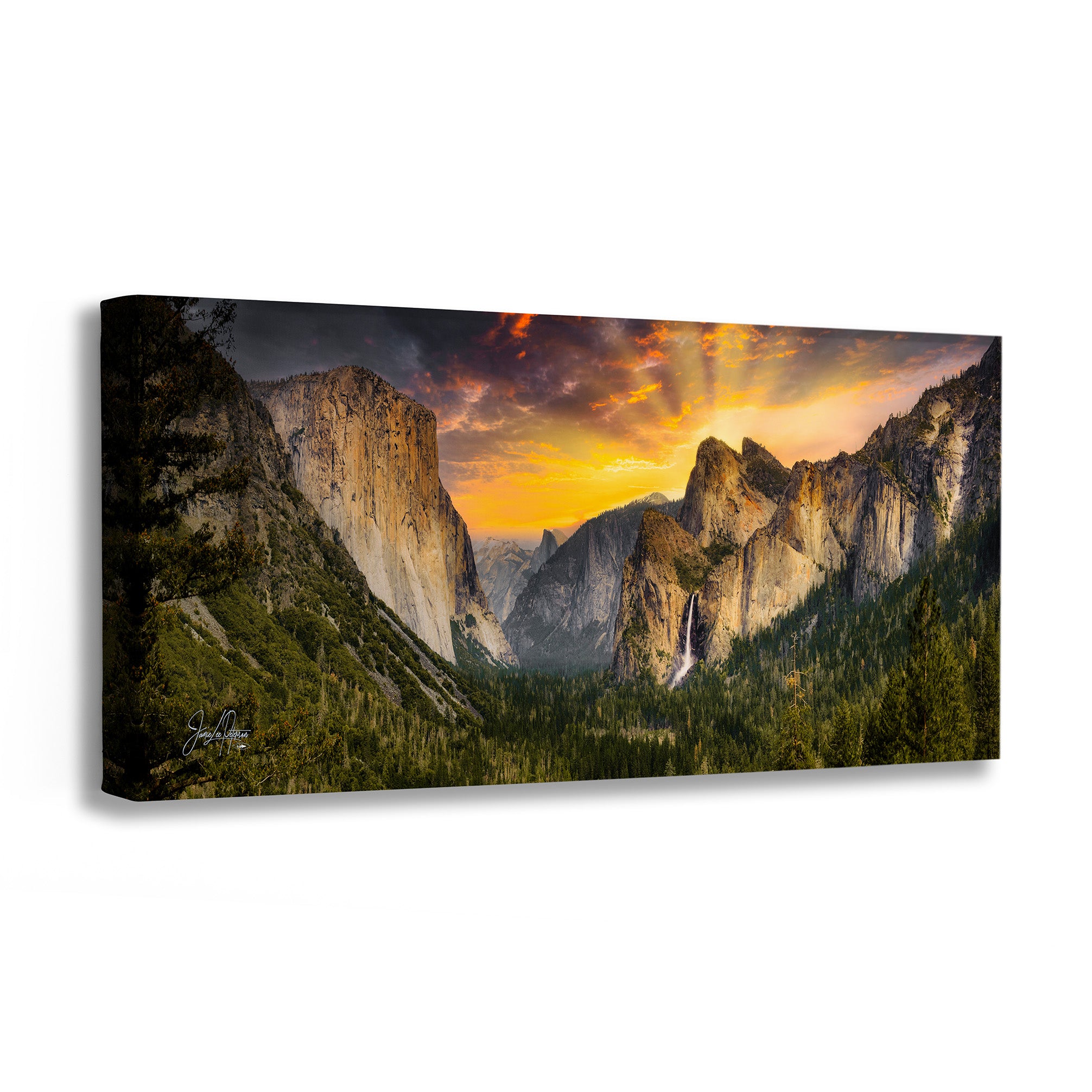 A rectangular canvas wall art print showing a dramatic mountain valley landscape at sunset, photographed against a plain white background. The artwork depicts tall rocky cliffs, dark green forested slopes, and a glowing orange and yellow sky with sun rays breaking through clouds.
