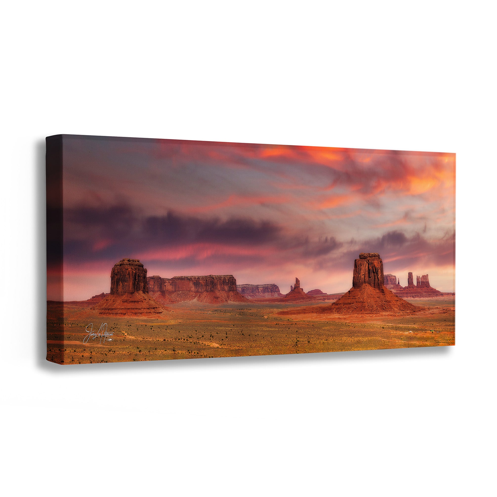 A rectangular canvas wall art print showing a desert landscape with large rock buttes under a dramatic orange, pink, and purple sunset sky, photographed against a plain white background.