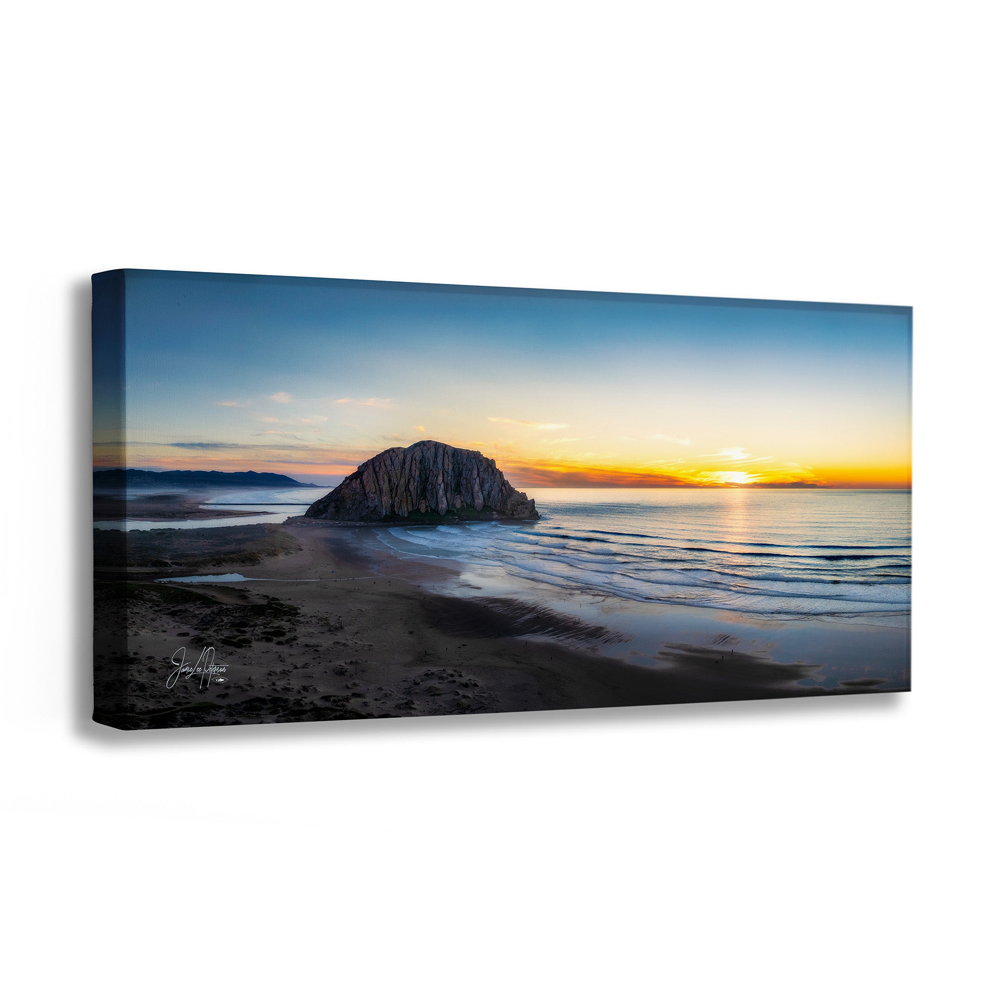 A panoramic canvas wall art print showing a coastal sunset scene with a large rock formation, calm water, and a glowing sun on the horizon, photographed against a light gray background.