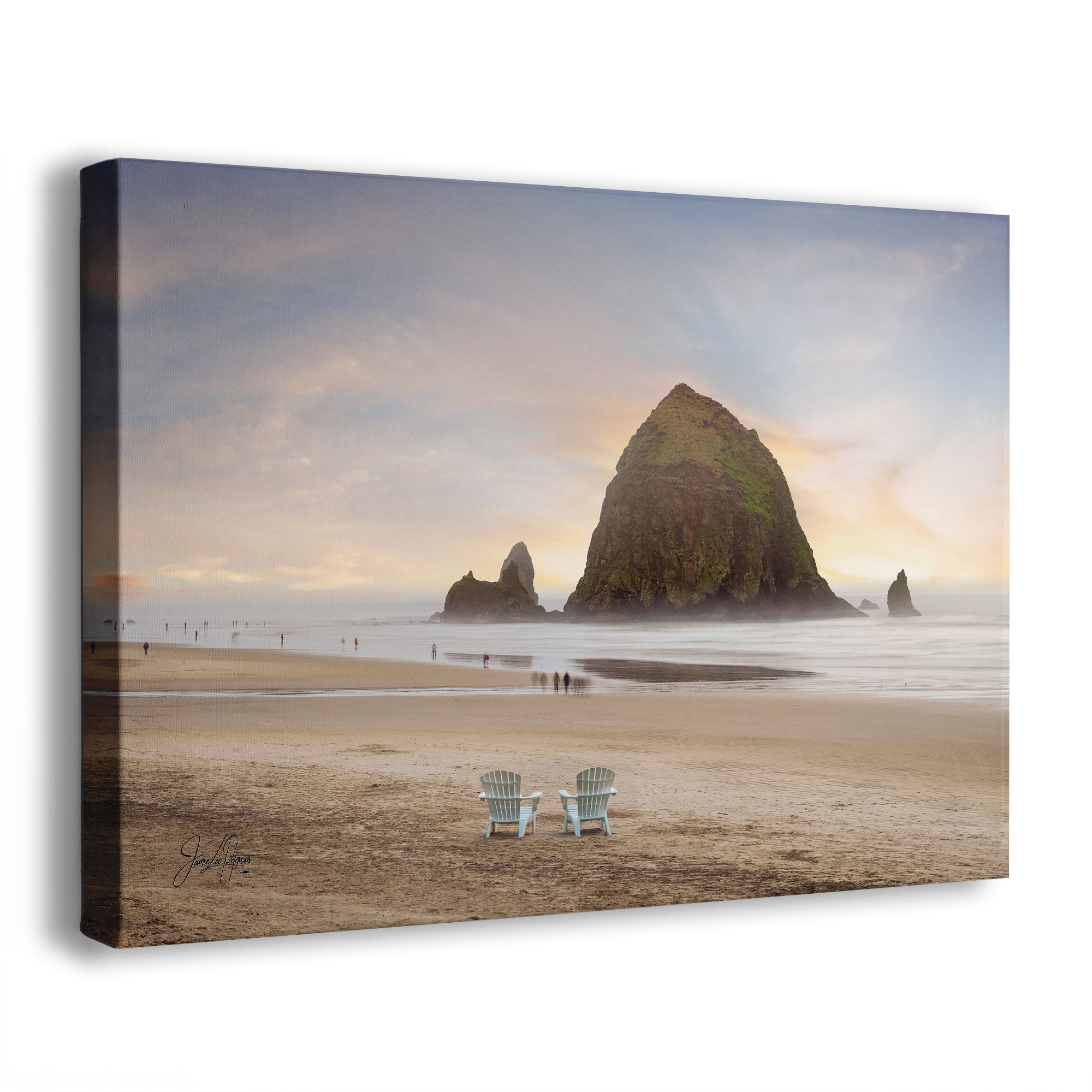 A canvas wall art print showing a beach scene with two white Adirondack chairs in the foreground, a large sea stack at Cannon Beach, Oregon, and smaller rocks in the water, and a soft sky with warm light near the horizon. The canvas is photographed at a slight angle on a plain white background.