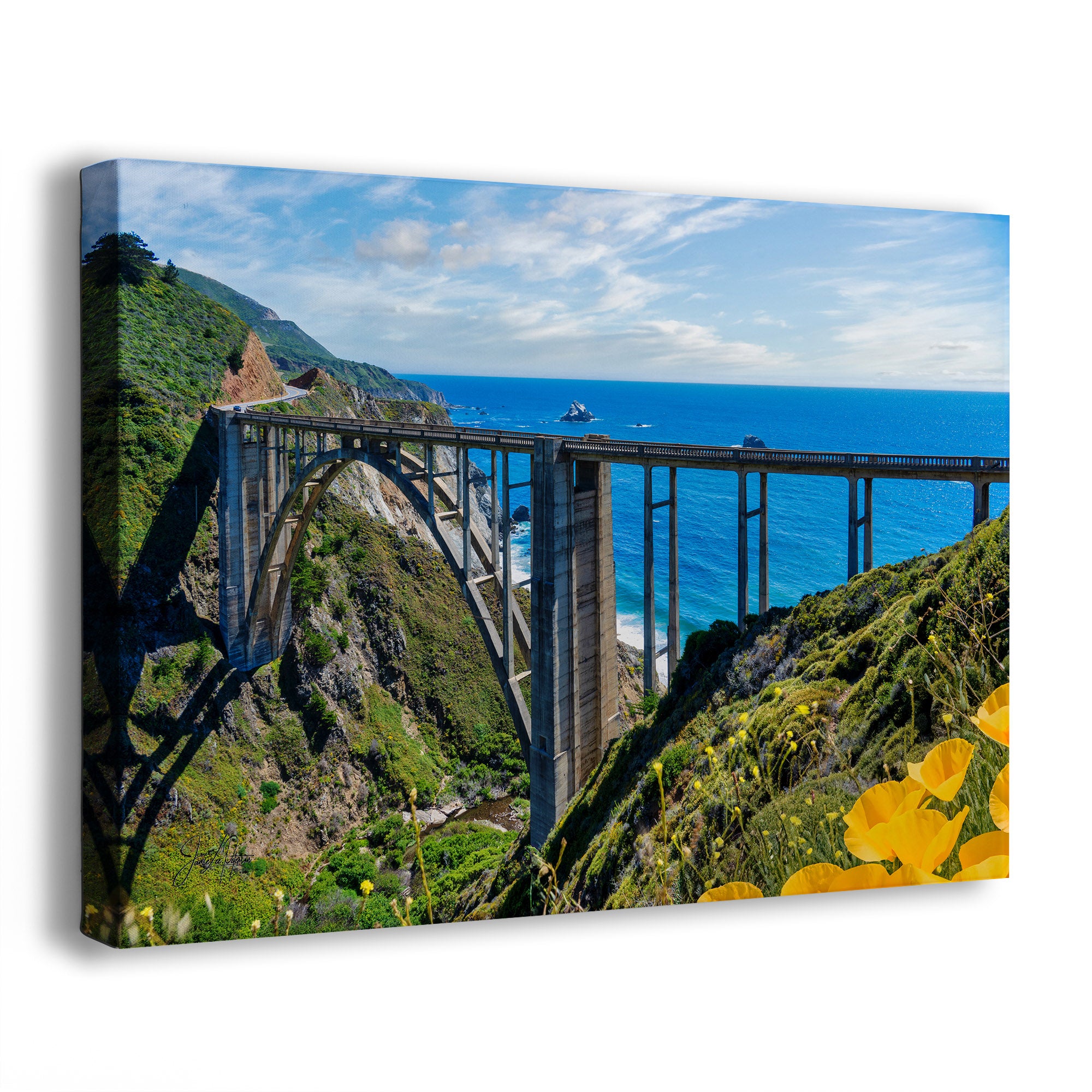 A canvas wall art print showing the historic Bixby Bridge spanning a deep canyon in Big Sur, California, with blue ocean water, green cliffs, and bright yellow poppies in the foreground, photographed against a plain white background.