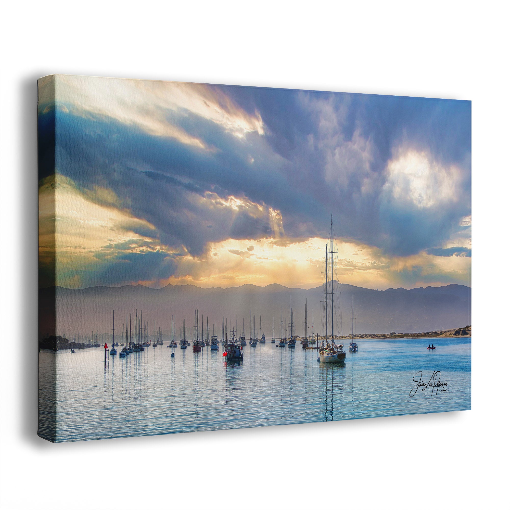 A canvas wall art print showing a harbor scene with sailboats on calm blue water, distant mountains, and dramatic clouds with sunlight breaking through. The canvas is photographed at a slight angle against a plain white background.