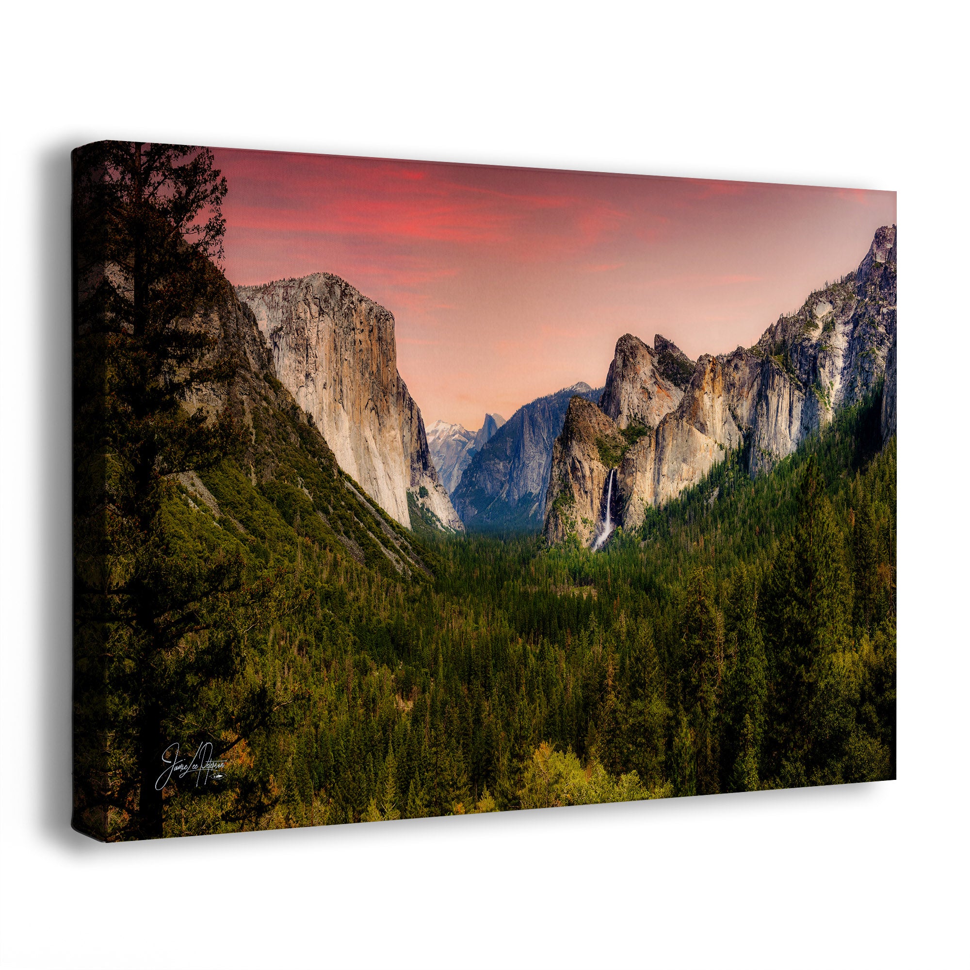 A rectangular canvas wall art print showing a mountain valley landscape at sunset, with tall rock cliffs, dense evergreen forest, and a pink-orange sky. The canvas is photographed at an angle against a plain white background.