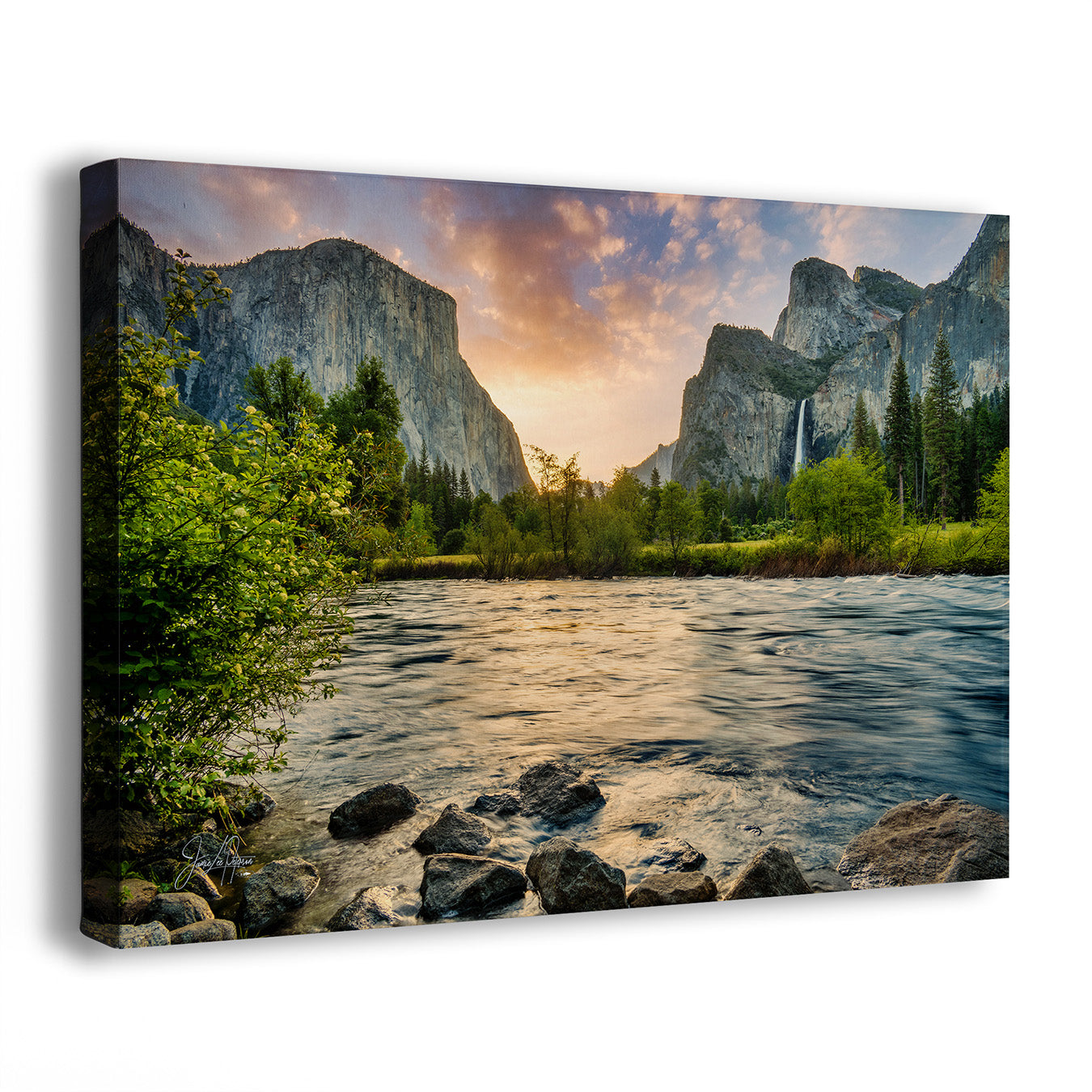 A rectangular canvas wall art print showing a mountain valley landscape with a river in the foreground, trees, cliffs, and a dramatic sunset sky. The canvas is photographed at a slight angle against a plain white background.