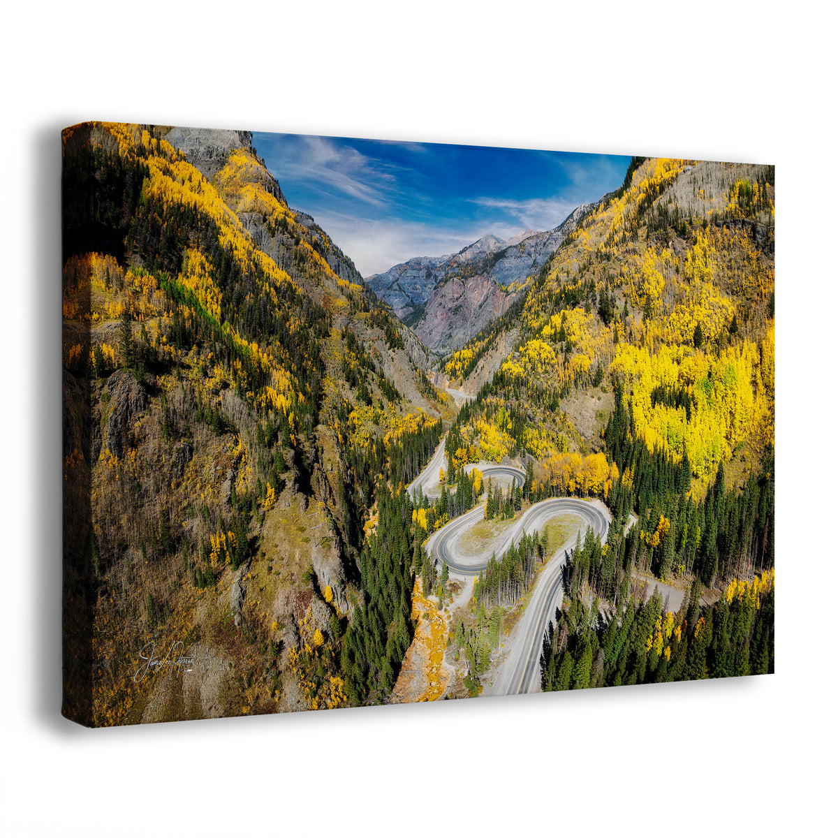 A canvas wall art print showing an aerial view Colorado's Million Dollar Highway, with bright yellow autumn foliage, evergreen trees, rocky cliffs, and a blue sky. The canvas is photographed at an angle against a plain white background.
