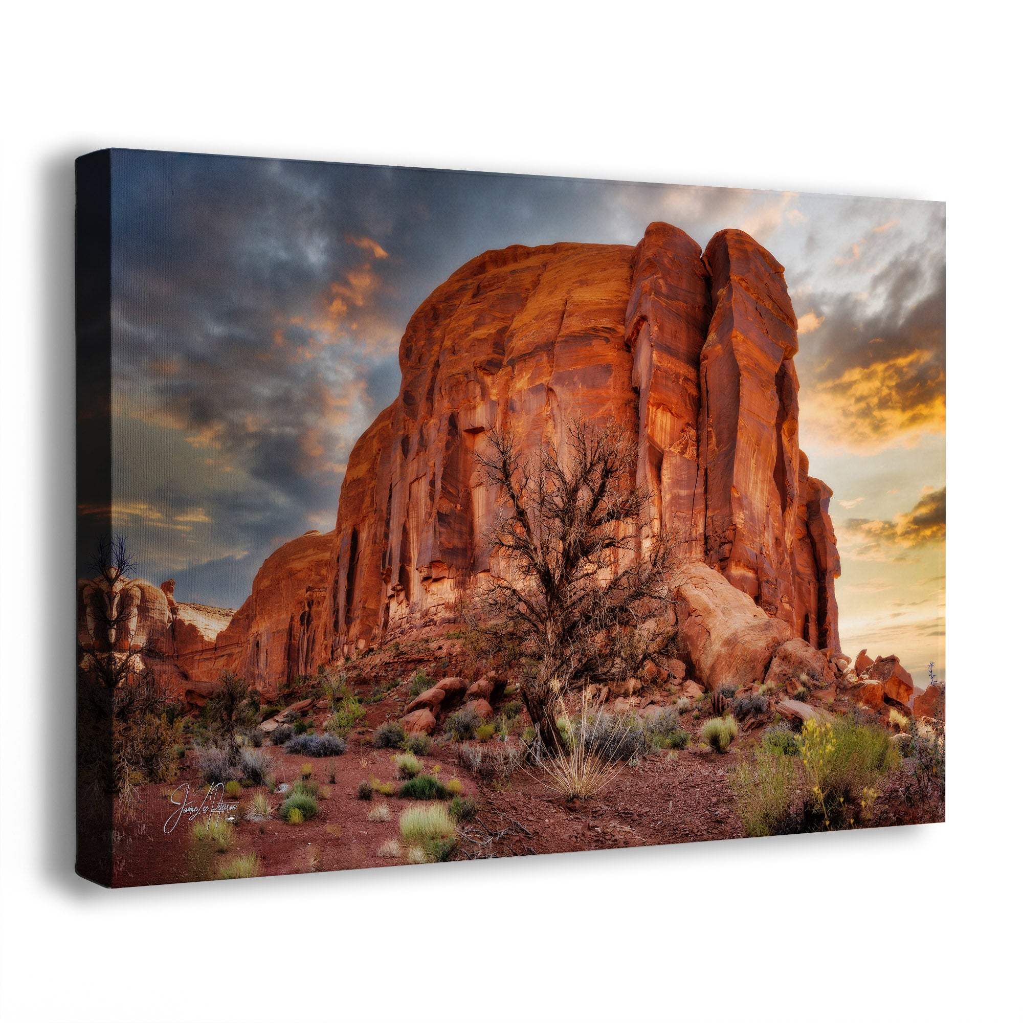 A rectangular canvas wall art print showing a desert rock formation at sunset, photographed against a plain white background. The artwork depicts orange and red sandstone cliffs with dark clouds and warm golden light in the sky.