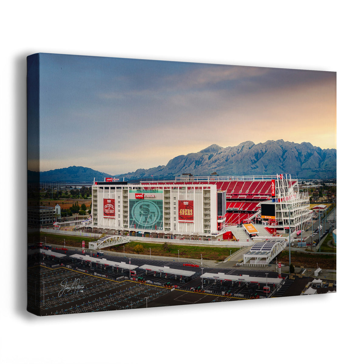 A Levi's Stadium sunrise wall art print showing an aerial view of the 49ers stadium with red seating, white structures, parking areas, and mountains in the background, all set against a cloudy sky. The canvas is photographed at an angle against a plain white background.