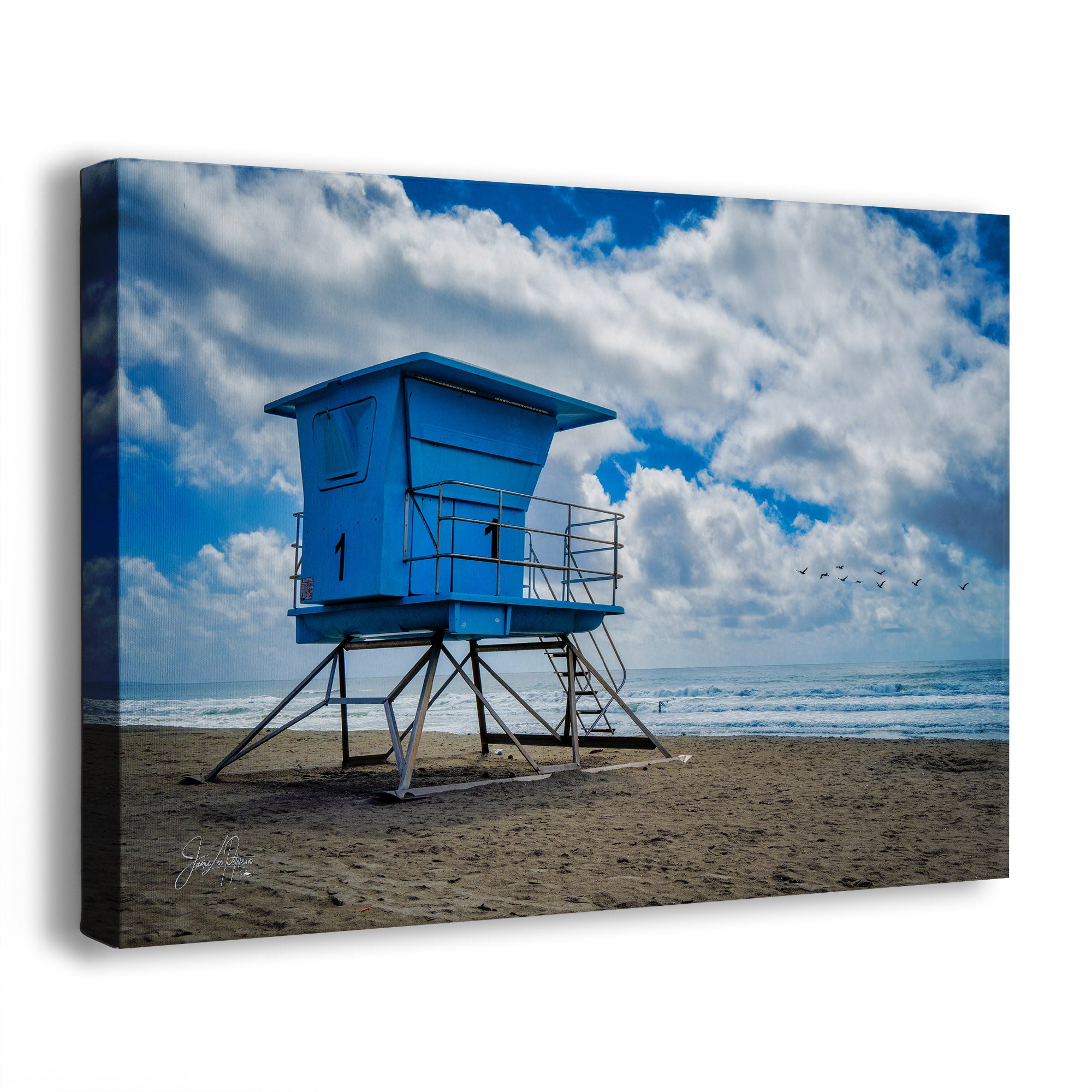A square canvas wall art print depicting a bright blue lifeguard tower on a sandy beach under a dramatic blue sky with large white clouds. The canvas is shown at an angle with a visible side edge and drop shadow on a plain white background.