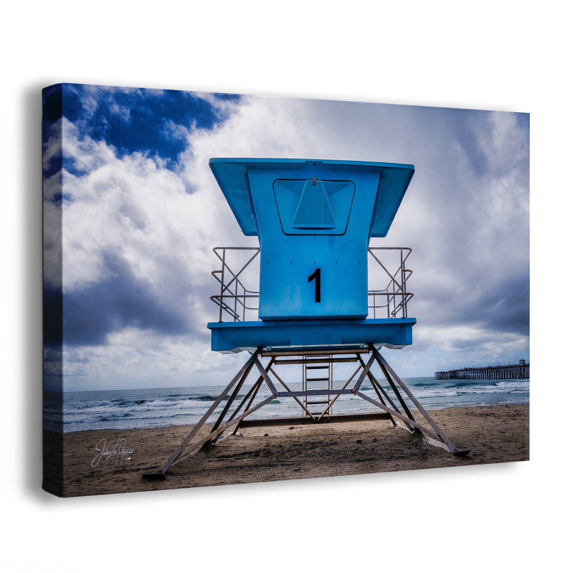 A canvas wall art print depicting a bright blue lifeguard tower on a sandy beach under a dramatic cloudy sky. The canvas is shown at a slight angle with a visible depth profile and shadow on a plain white background.