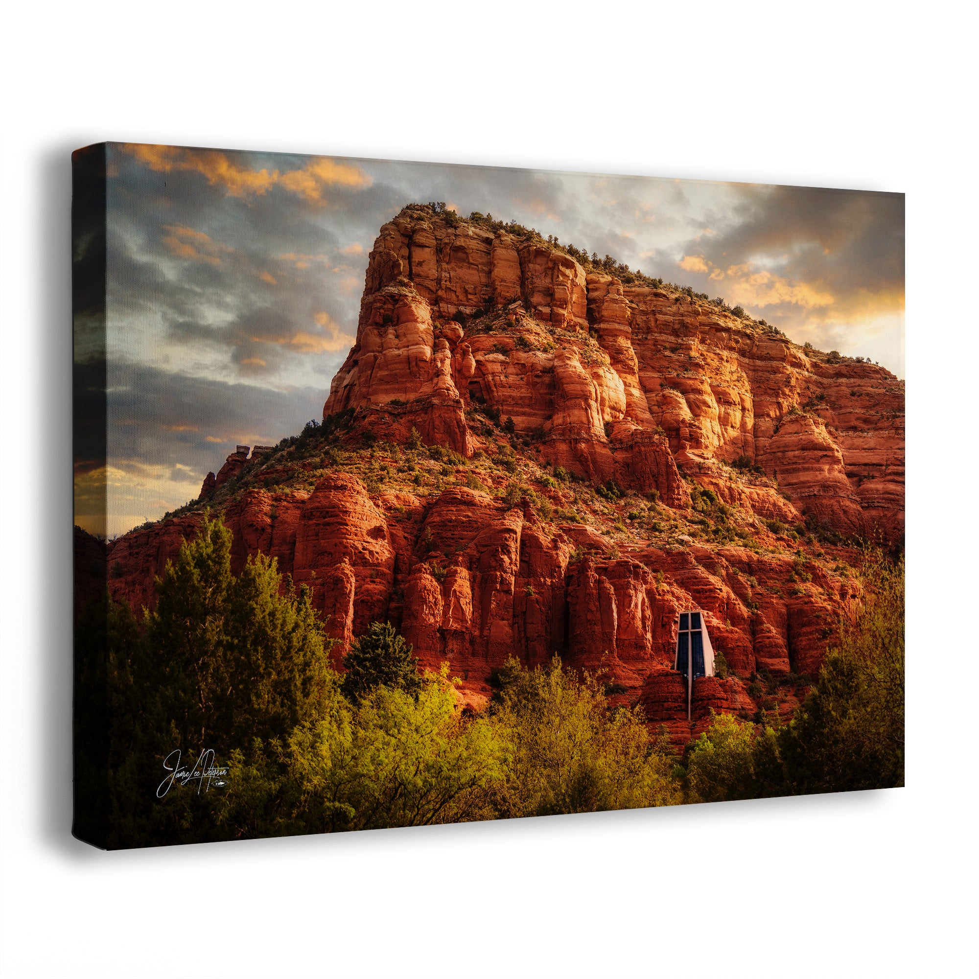 A canvas wall art print showing a desert rock formation in Sedona, Arizona, at sunrise, with the Chapel of the Holy Cross photographed on a clean white background. The artwork depicts orange-red sandstone cliffs, green trees in the foreground, and a dramatic cloudy sky with warm golden light.