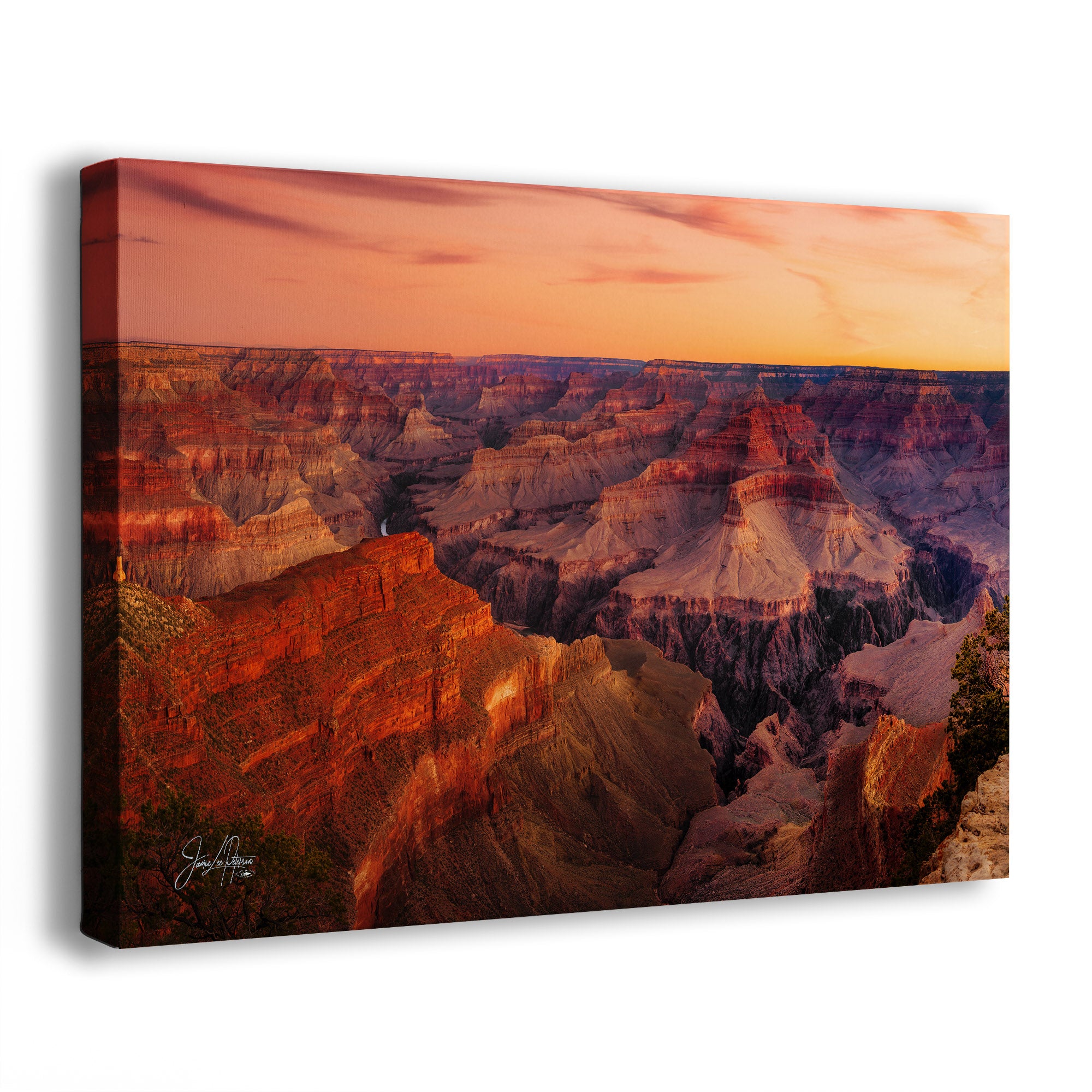 A wall art print showing a dramatic canyon landscape at sunset, photographed at an angle against a plain white background. The artwork depicts layered red, orange, brown, and purple rock formations under a warm peach sky.